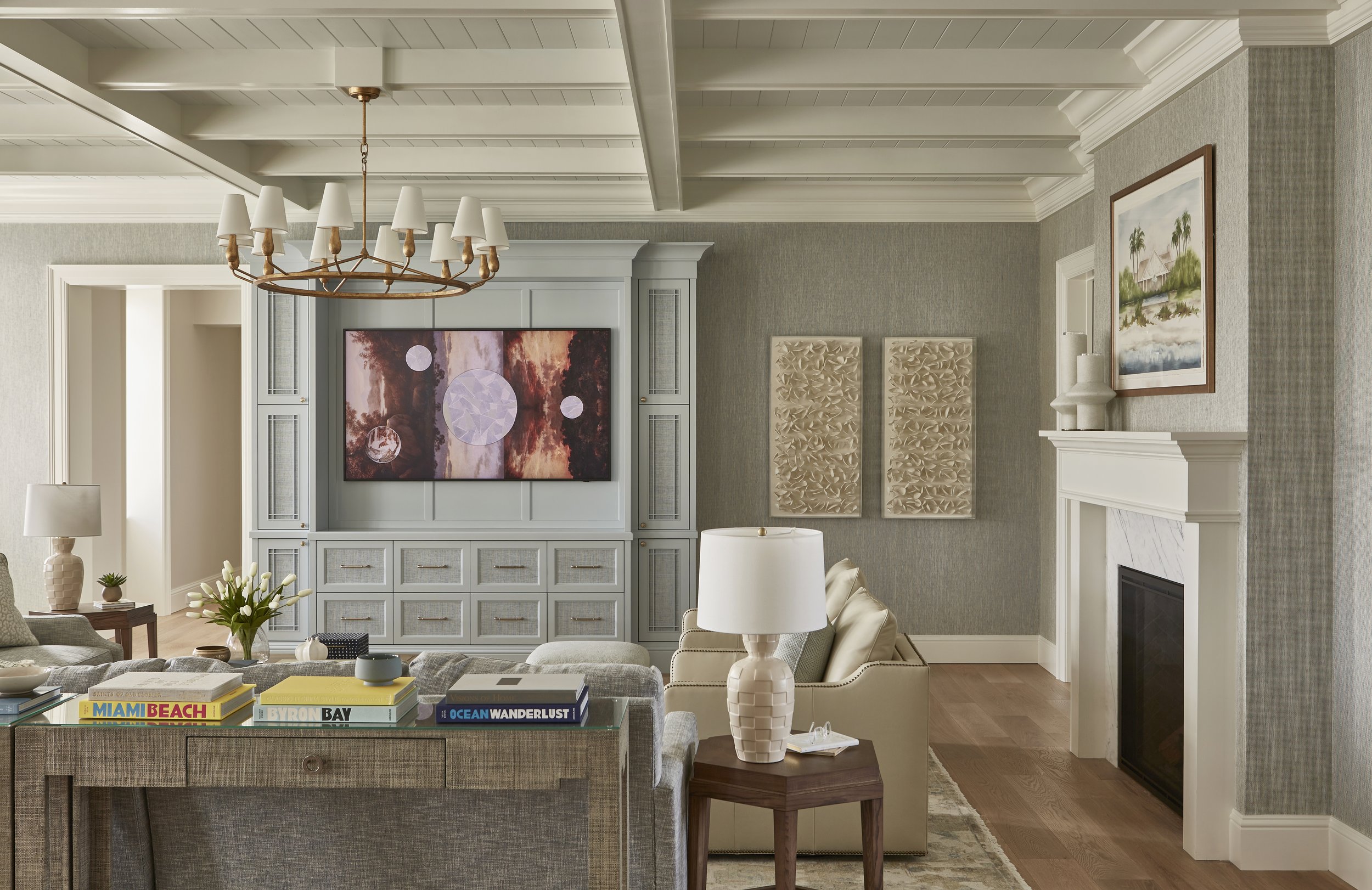 A cozy living room with a vaulted ceiling, white beams, and a chandelier. There is a built-in white entertainment unit with a large router on a TV screen displaying abstract art. The room has a white fireplace with a framed landscape painting above i