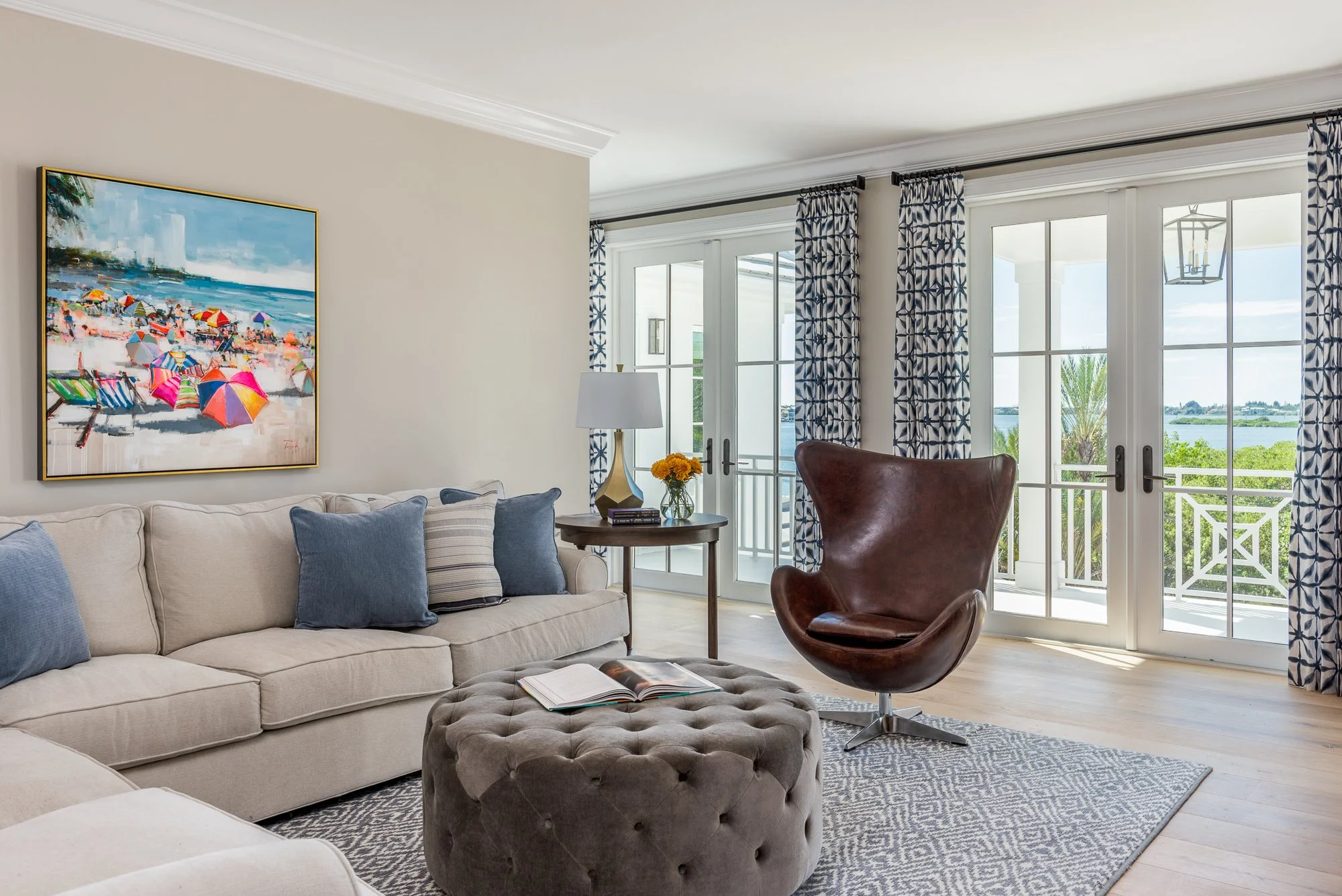 Living room with beige sofa, blue and striped pillows, round gray tufted ottoman with open book, brown leather swivel chair, side table with lamp and yellow flowers, large windows with blue patterned curtains, view of water and greenery outside.