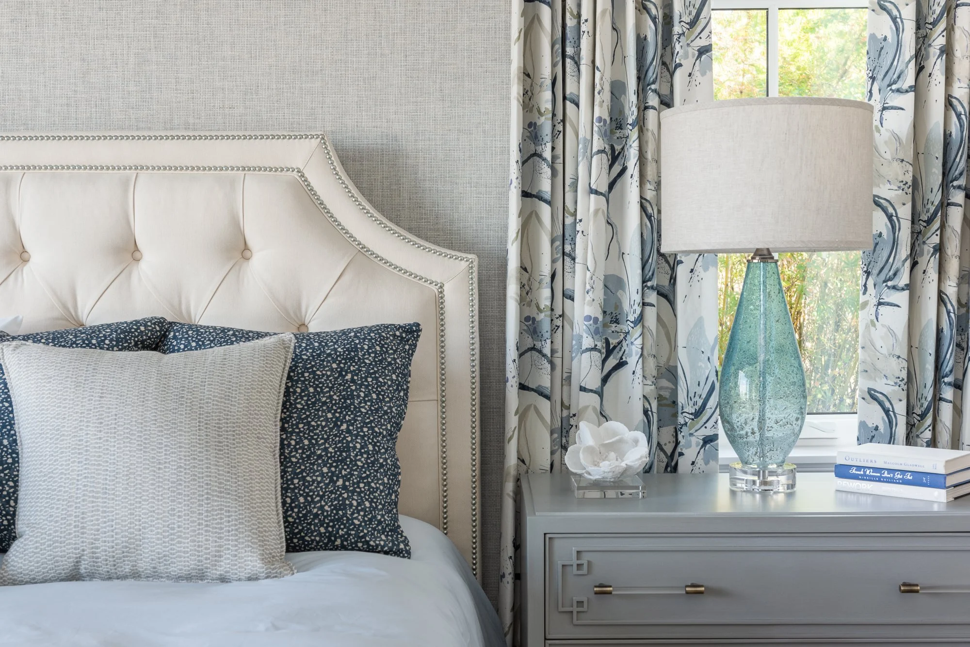 A bedroom with a beige tufted headboard, blue and white pillows, a gray nightstand with a decorative white shell and a framed white object, a glass table lamp with a beige shade, and a window with floral curtains showing greenery outside.