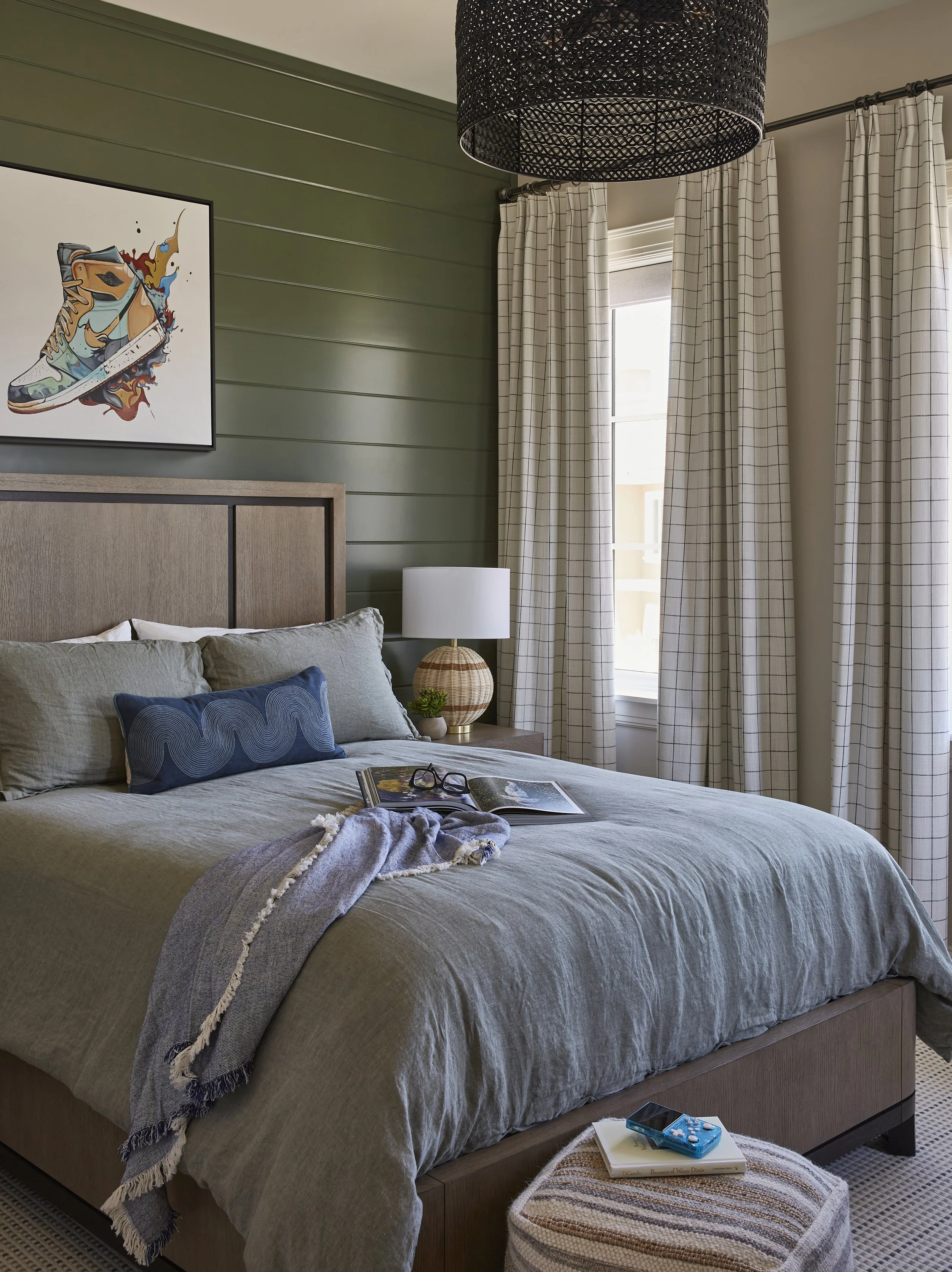 A bedroom with a bed against a green wall, decorated with a large pillow and a blanket, a bedside table with a lamp, a pair of glasses, an open magazine, and a tray with a book and a game. The window has checkered curtains, and there is a black penda