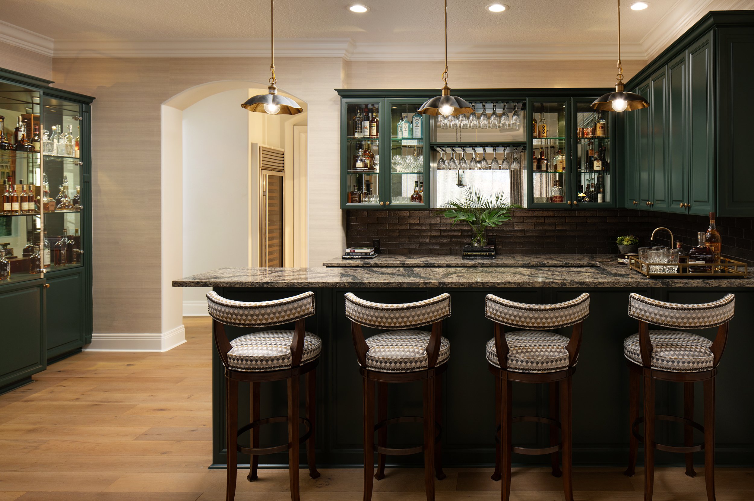 Kitchen bar area with dark green cabinets, granite countertop, hanging pendant lights, barstools with patterned cushions, and a display of bottles and glasses.