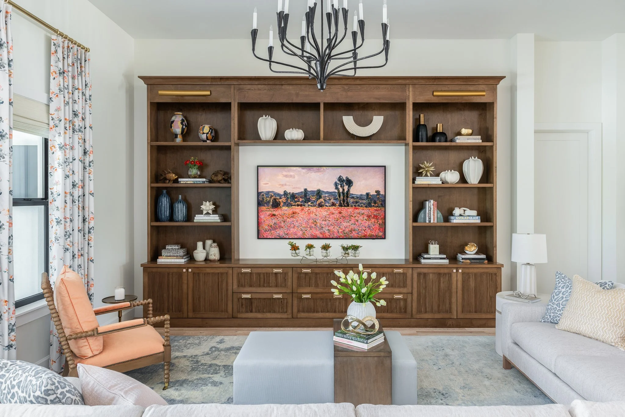 Living room with a built-in wooden bookshelf, a framed landscape painting, a round vase with white flowers on the coffee table, and seating including a peach armchair and a white sofa with pillows.