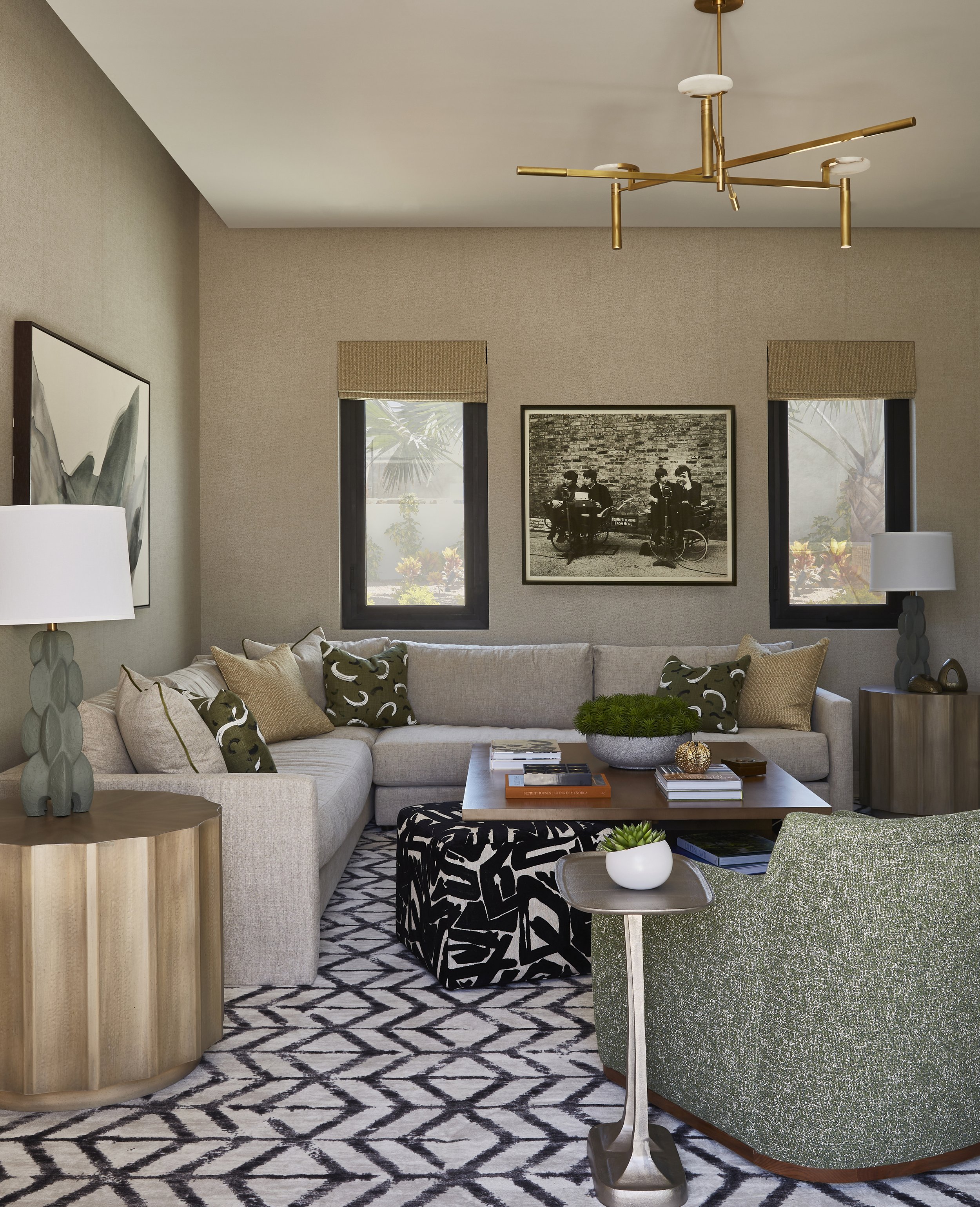 A living room with a beige sectional sofa, green and beige throw pillows, two side tables with lamps, a black-and-white patterned ottoman, a wooden coffee table with books and decorations, a green armchair, and two windows with beige blinds and a bla