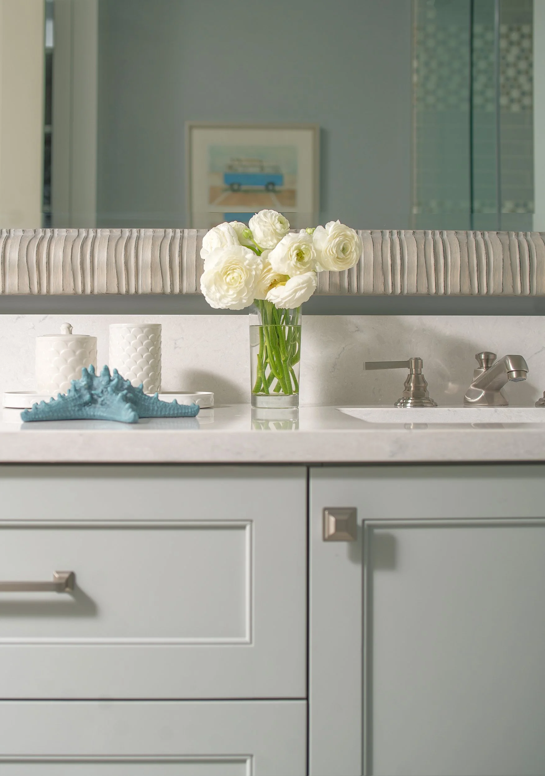 White flowers in a glass vase on a white bathroom countertop with decorative items, reflected in a mirror above, with gray cabinetry and a framed picture in the background.