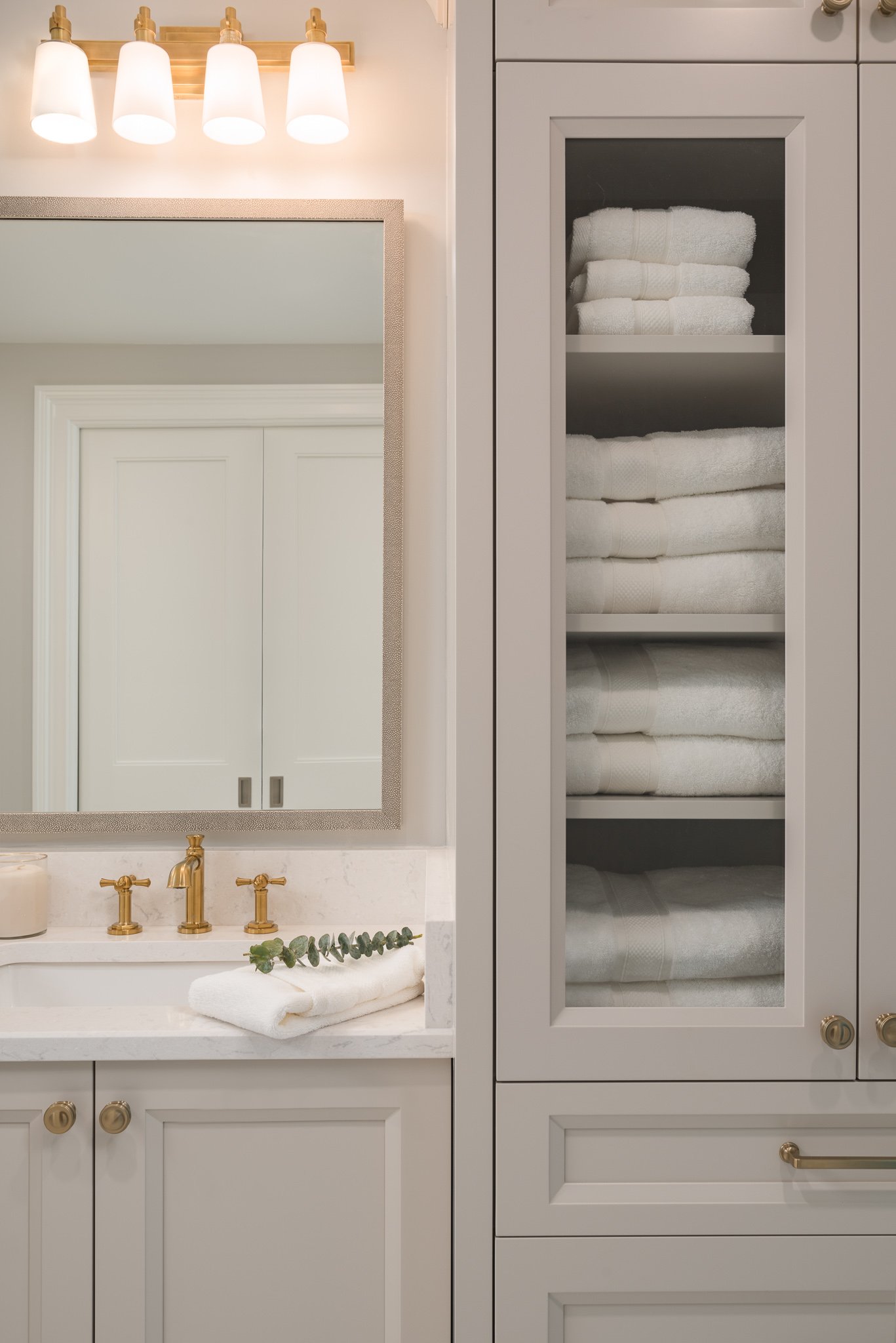 Bathroom cabinet with neatly folded white towels on shelves, gold hardware, and a mirror reflecting white double doors.