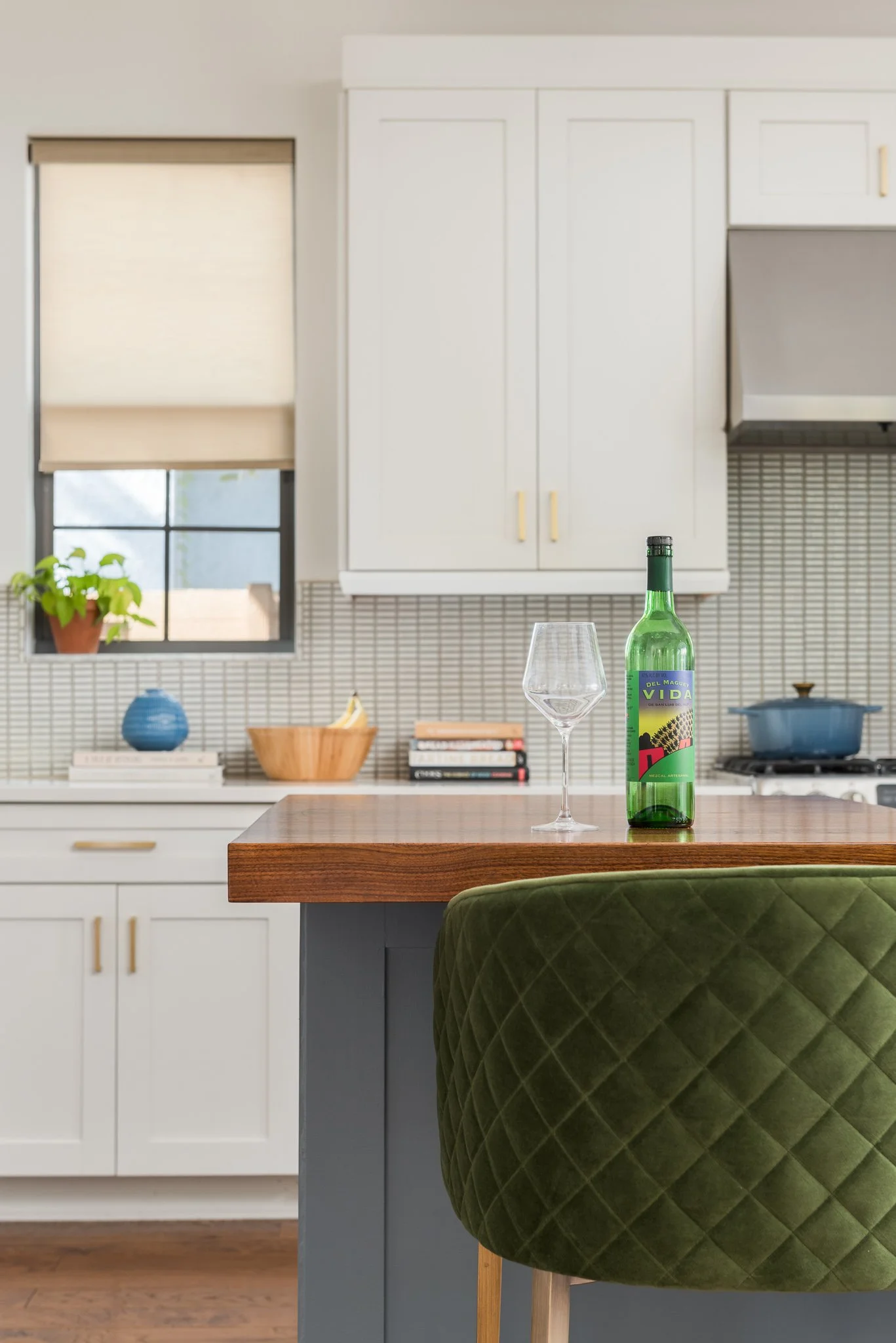 Kitchen with a wooden island, wine bottle, and empty wine glass on the counter. Green upholstered chair partially visible in the foreground. White cabinets, small window with blinds, and decorative items on the backsplash.