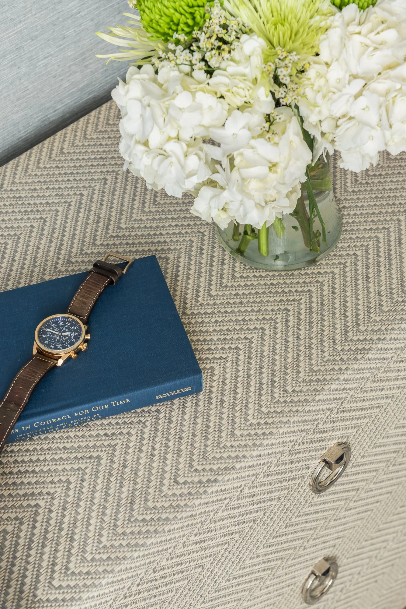 A glass vase with white and green flowers on a covered dresser with two drawers, a blue book, and a wristwatch on top.