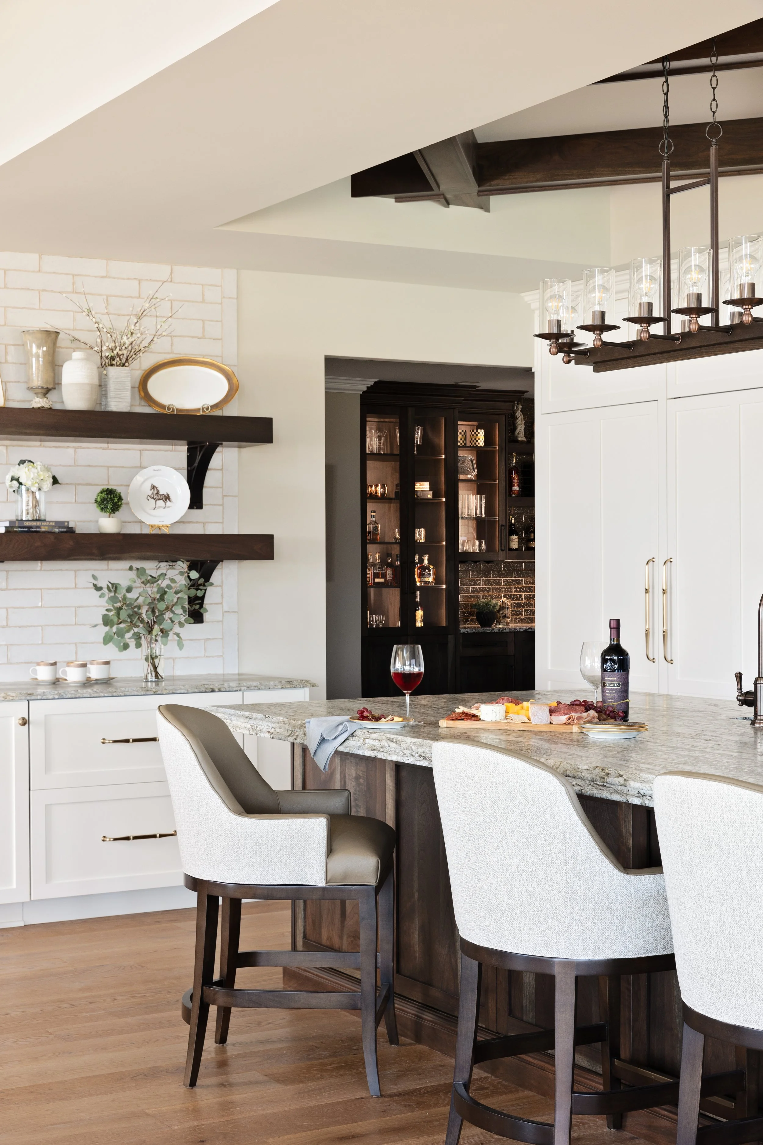 Modern kitchen with white and dark wood cabinetry, granite countertops, a kitchen island, and bar stools. There is a wine glass and a bottle of wine on the counter, and a view into a bar area with glass shelves and liquor bottles.