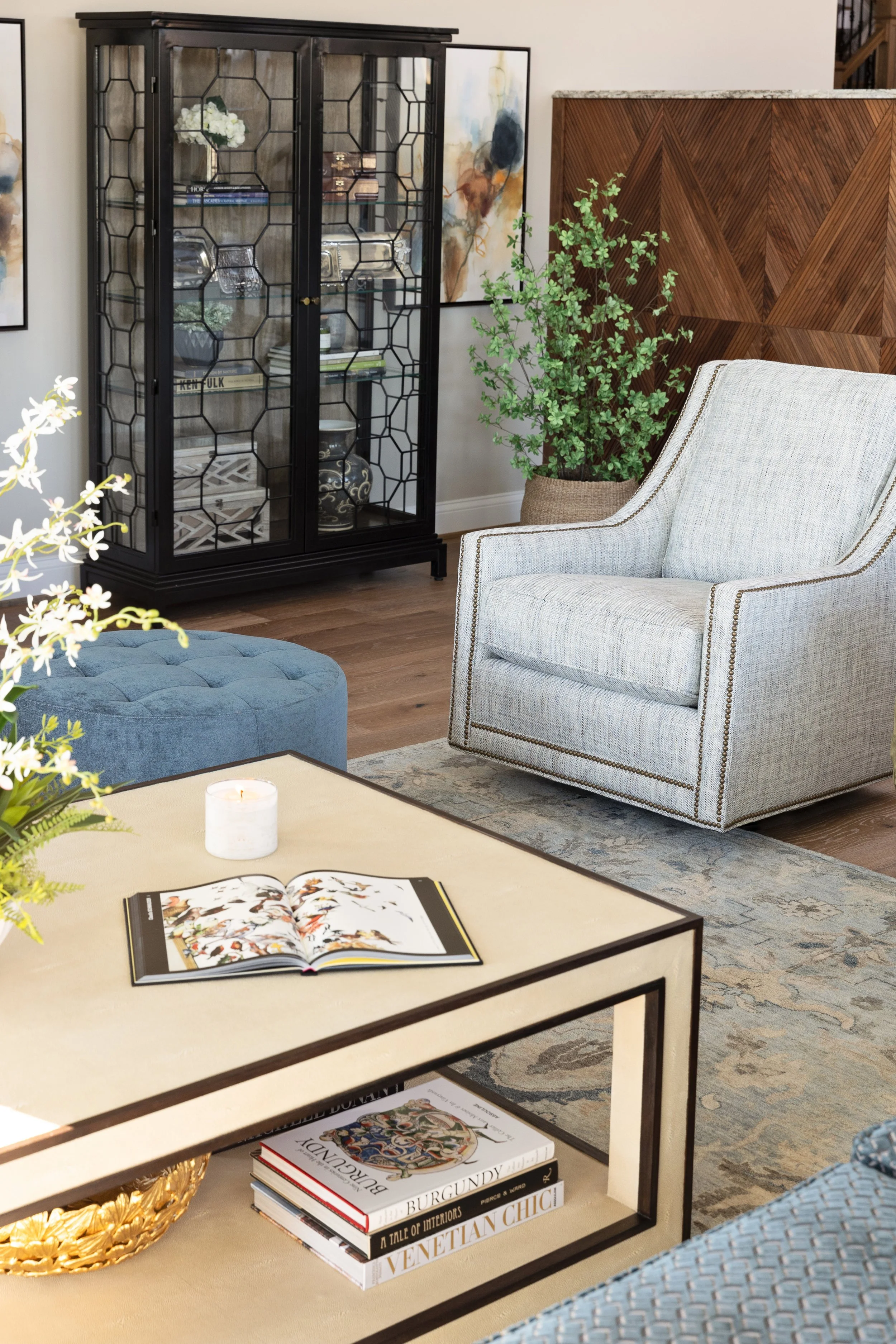 Living room with a white and gray armchair, a blue tufted ottoman, a beige coffee table with books and a candle, a black glass cabinet, and potted plants, with abstract art on the wall.