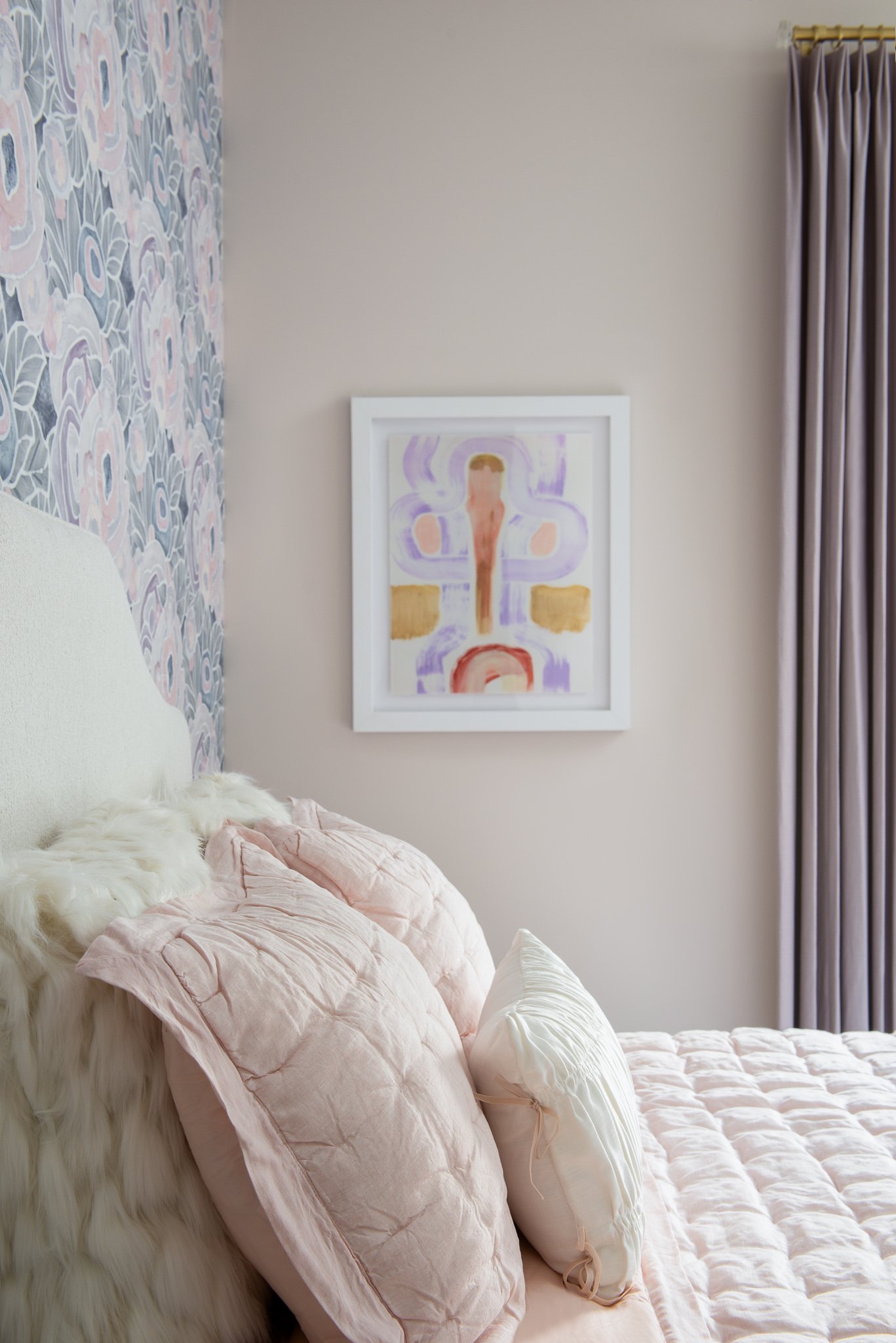 A bed with light pink and white pillows, a fluffy white throw, and a quilted pink bedspread, positioned against a wall with floral wallpaper on one side and a plain wall with an abstract art piece on the other. Cream curtain hanging on the window.