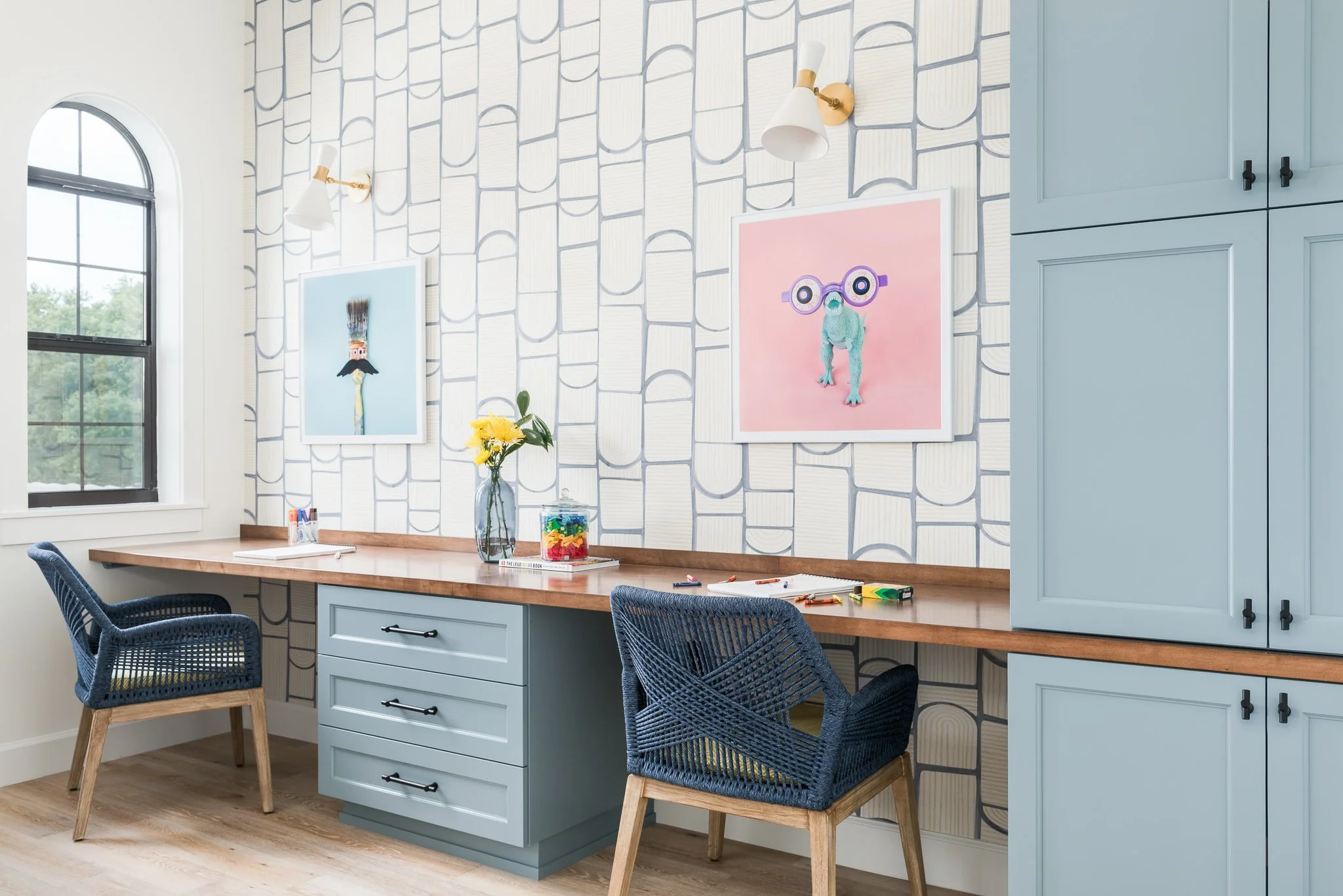 A home office or craft room with a wooden desk, blue drawers, and matching cabinets. Two blue woven chairs are positioned at the desk. The room has a window with black framing and a tiled patterned wallpaper with books motifs. There are two wall-moun