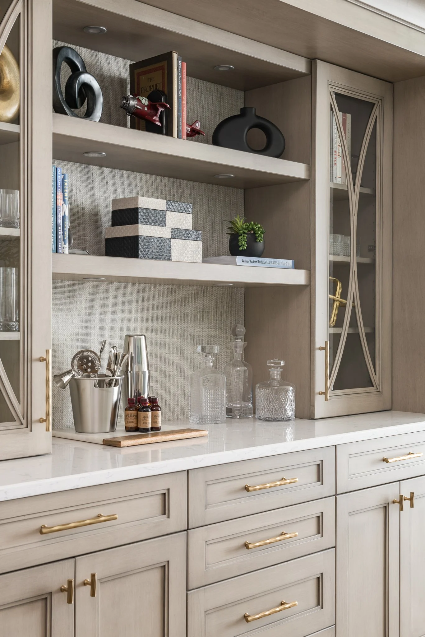 A kitchen cabinet with a marble countertop, displaying glass decanters, small bottles, and kitchen utensils. The upper shelves hold books, decorative boxes, and vases, with a small potted plant.