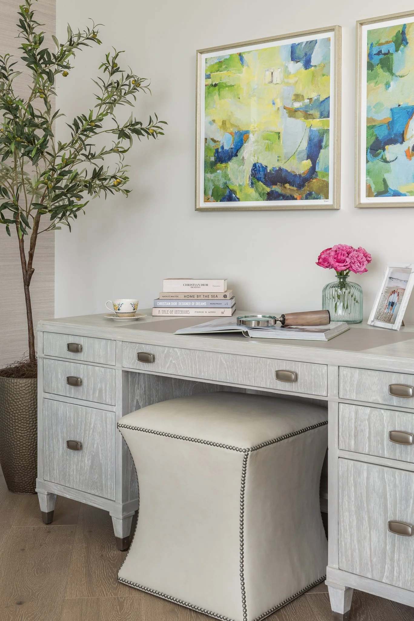 A white desk with a pile of books, a cup, a magnifying glass, a vase with pink flowers, a framed photo, and a large watercolor painting on the wall behind it. A tall potted plant is on the left.