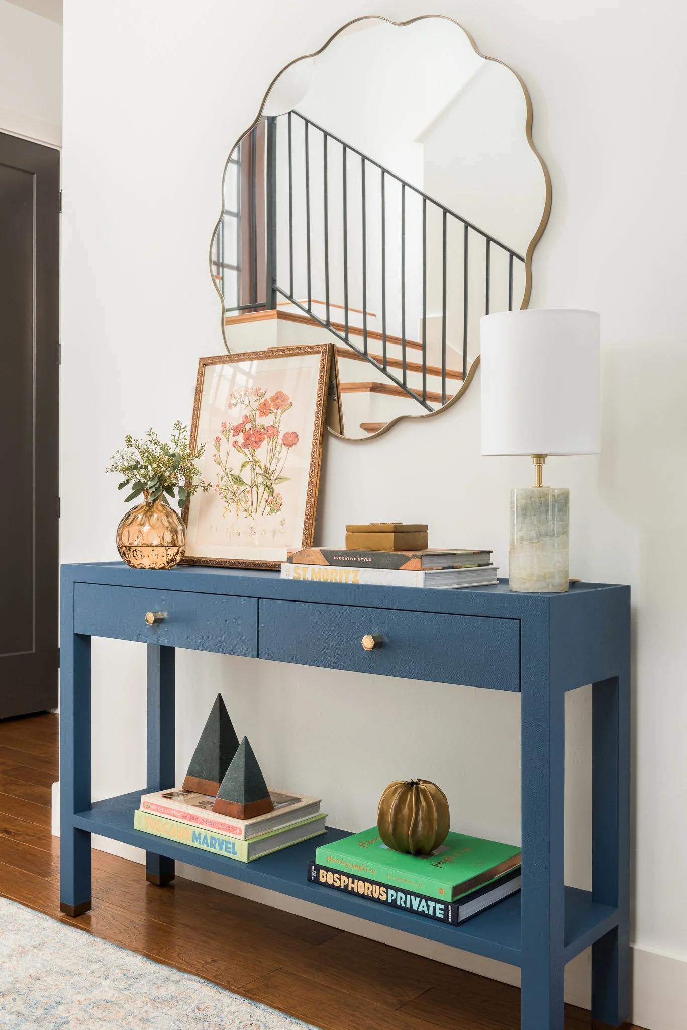 A blue console table with books, decorative objects, a table lamp, and a framed artwork, with a large, irregularly shaped mirror above it reflecting a staircase and railing.