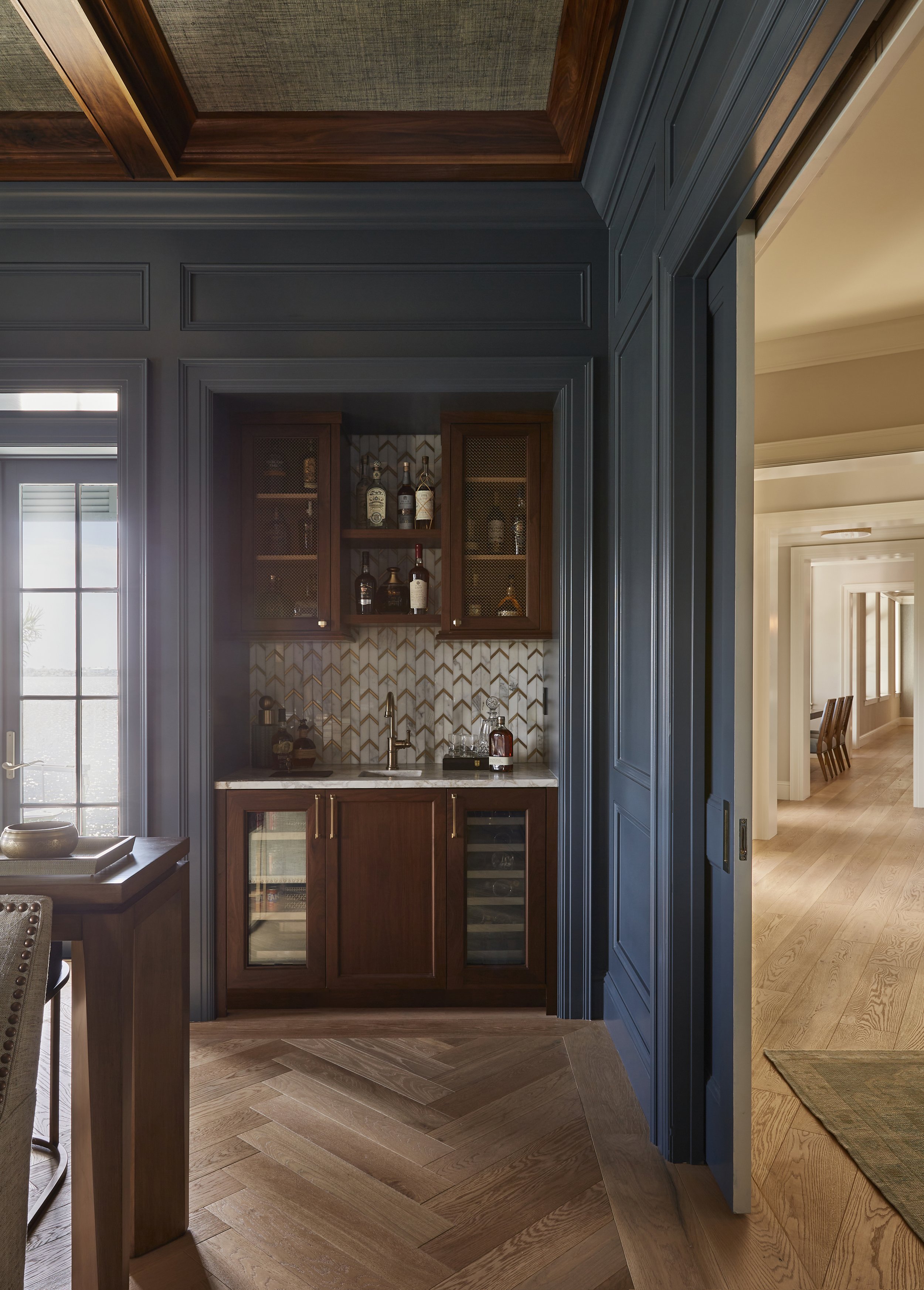 A home bar area with dark wood cabinetry, a marble countertop, a glass bottle collection, and a herringbone hardwood floor, adjacent to a hallway with lighter wood floors.