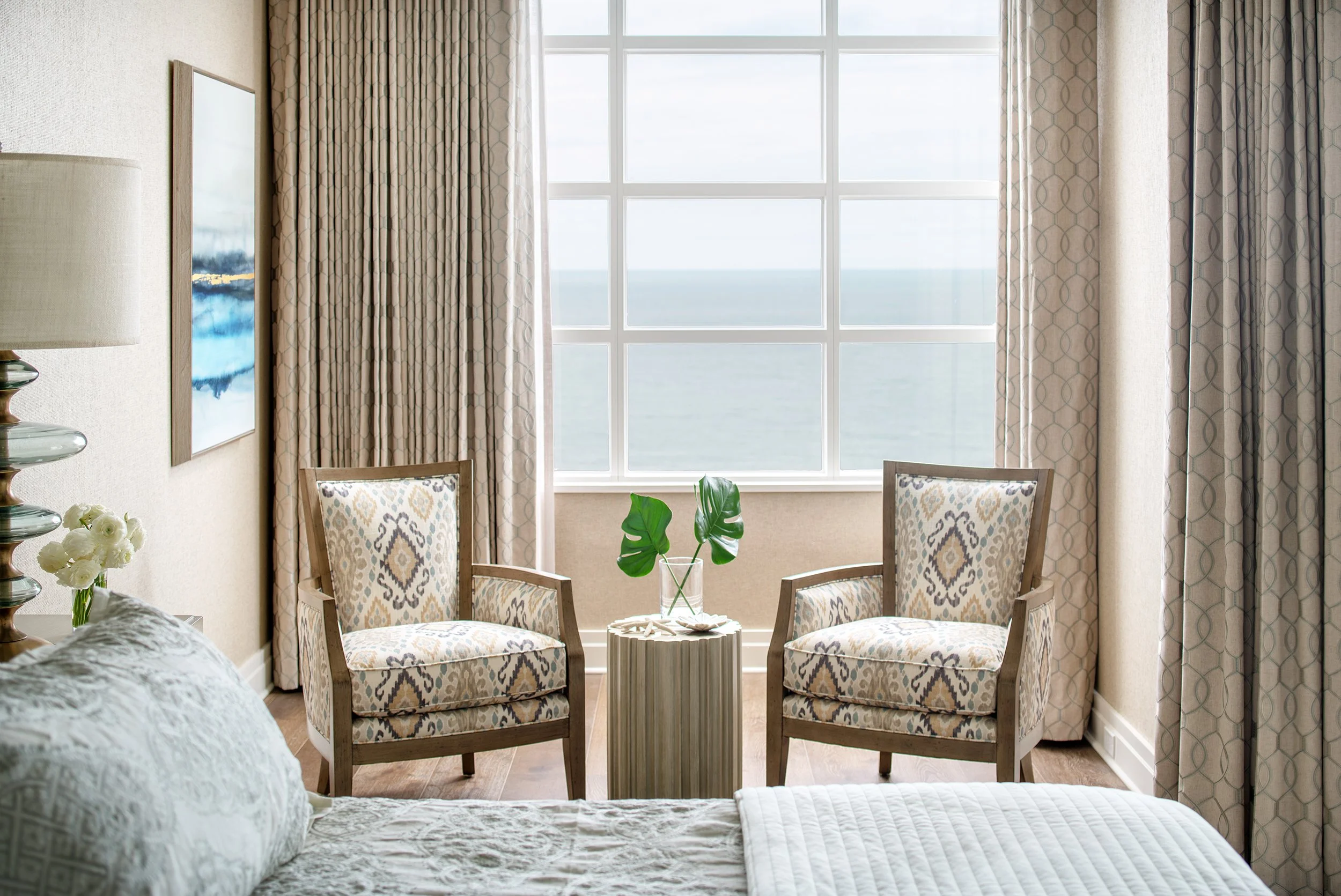 A cozy bedroom with two patterned armchairs facing a large window overlooking the ocean, with a small side table holding a vase with green leaves.