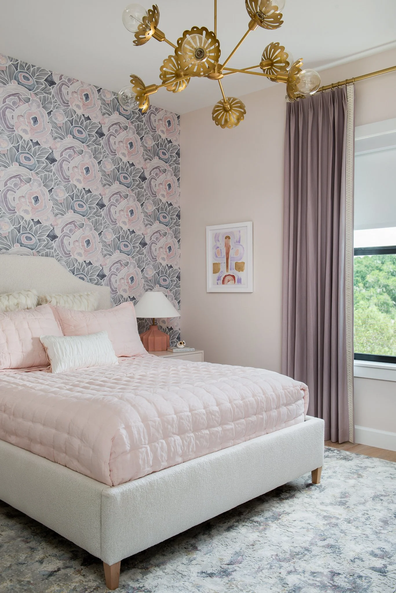 A bedroom with a pink quilted bed, pink pillows, a white headboard, a white nightstand with a pink lamp, purple curtains, a window with a view of trees, a modern chandelier with gold accents, an abstract art piece on the wall, and a floral wallpaper 