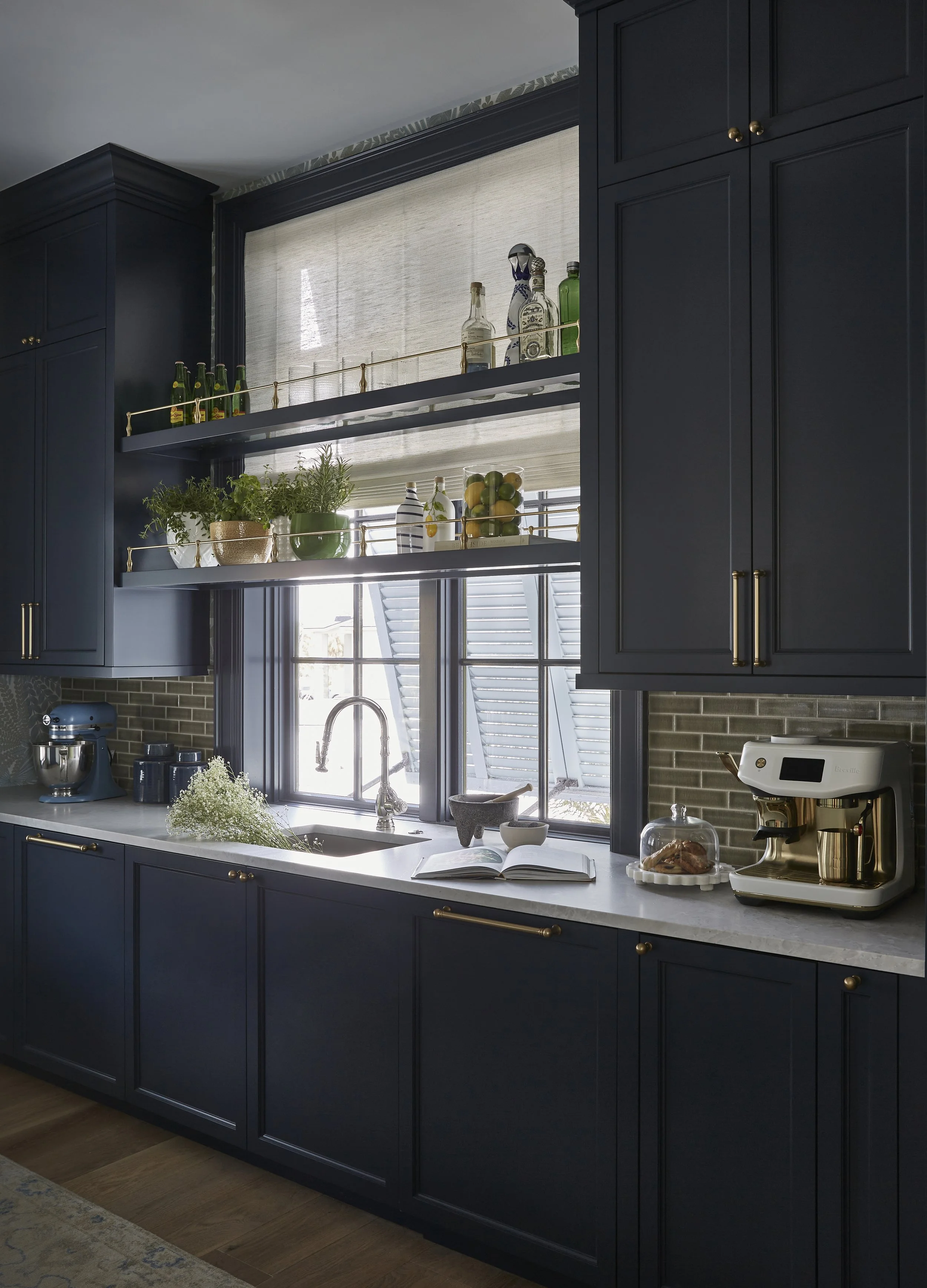 A kitchen with blue cabinets, open shelves above the sink with bottles and potted plants, a window behind the sink, a cookbook and coffee maker on the countertop.