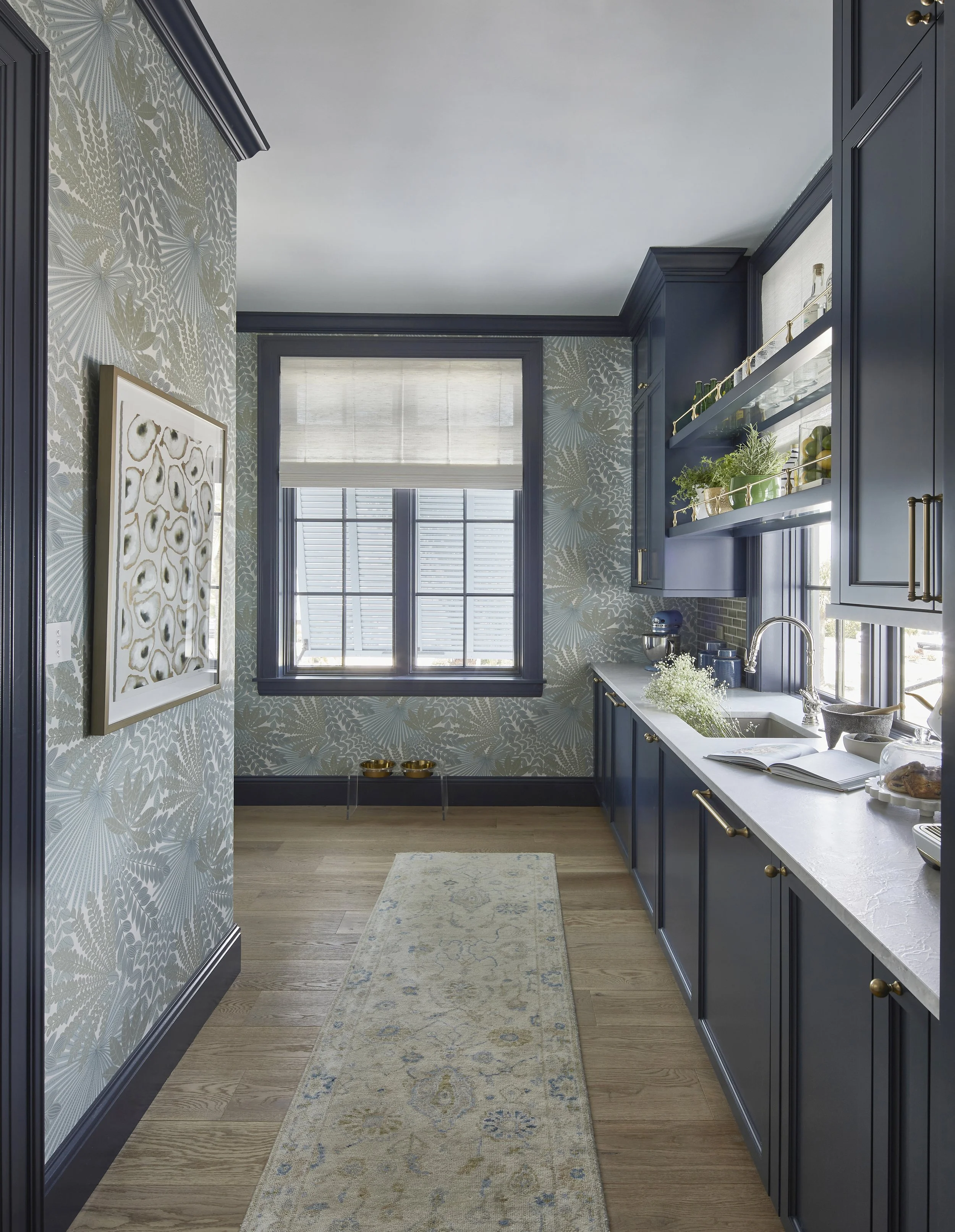 A bright kitchen with navy blue cabinets, gold hardware, patterned wallpaper, a white countertop, a window with blinds, and a beige patterned rug on wooden flooring.