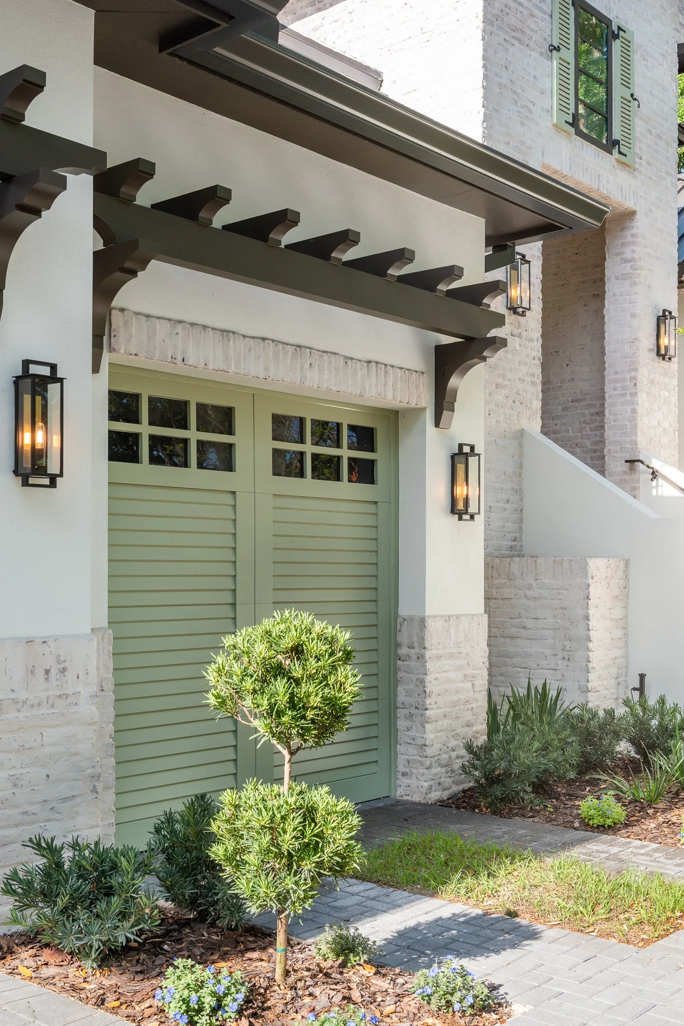 Exterior view of a modern house with white brick and stucco walls, a green garage door, outdoor wall lanterns, and landscaped bushes and grass.