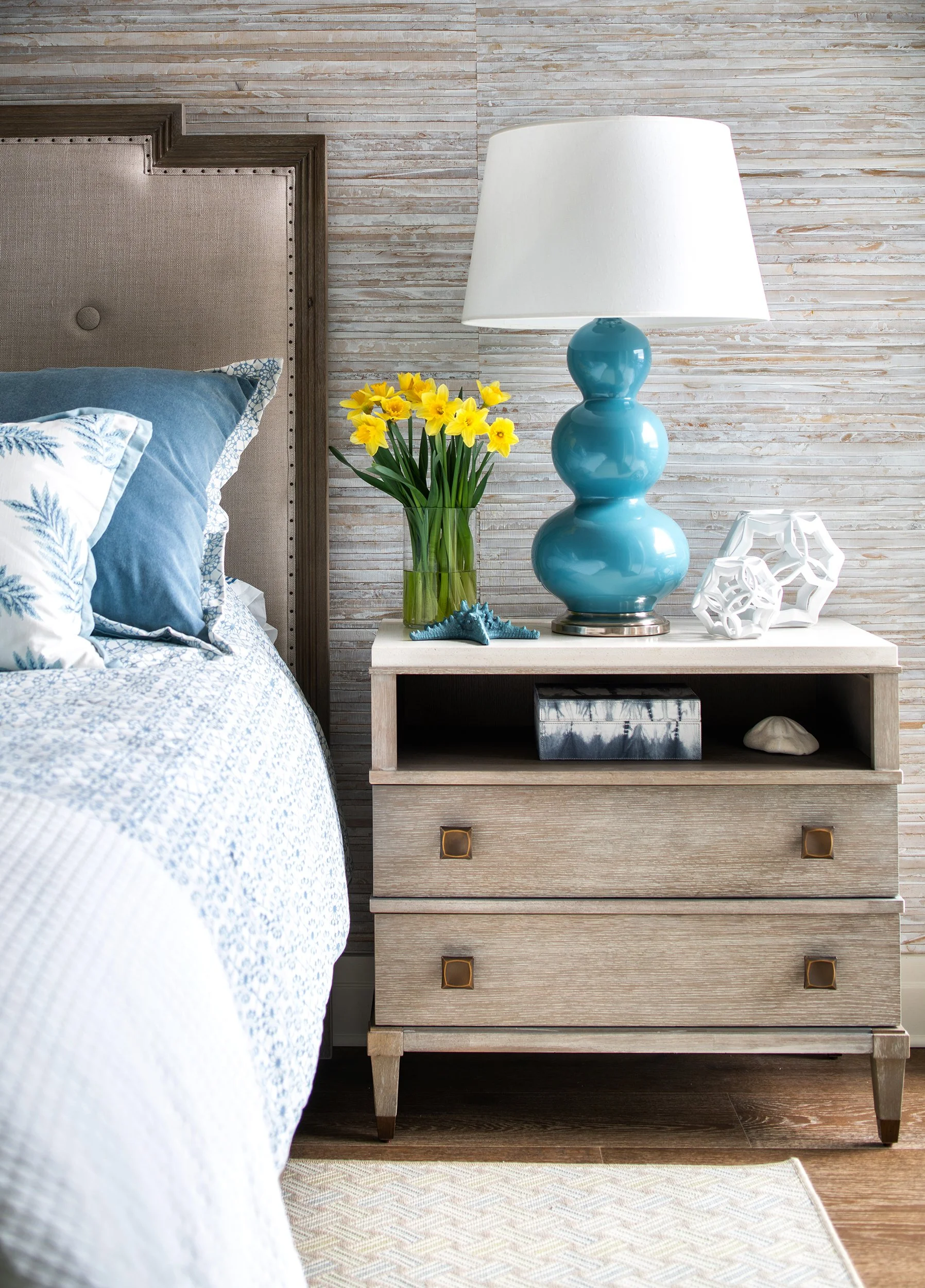 A bedroom nightstand with a large turquoise ceramic lamp, a clear vase of yellow daffodils, white decorative objects, and a black box, beside a bed with blue and white patterned bedding, against a textured beige wall.