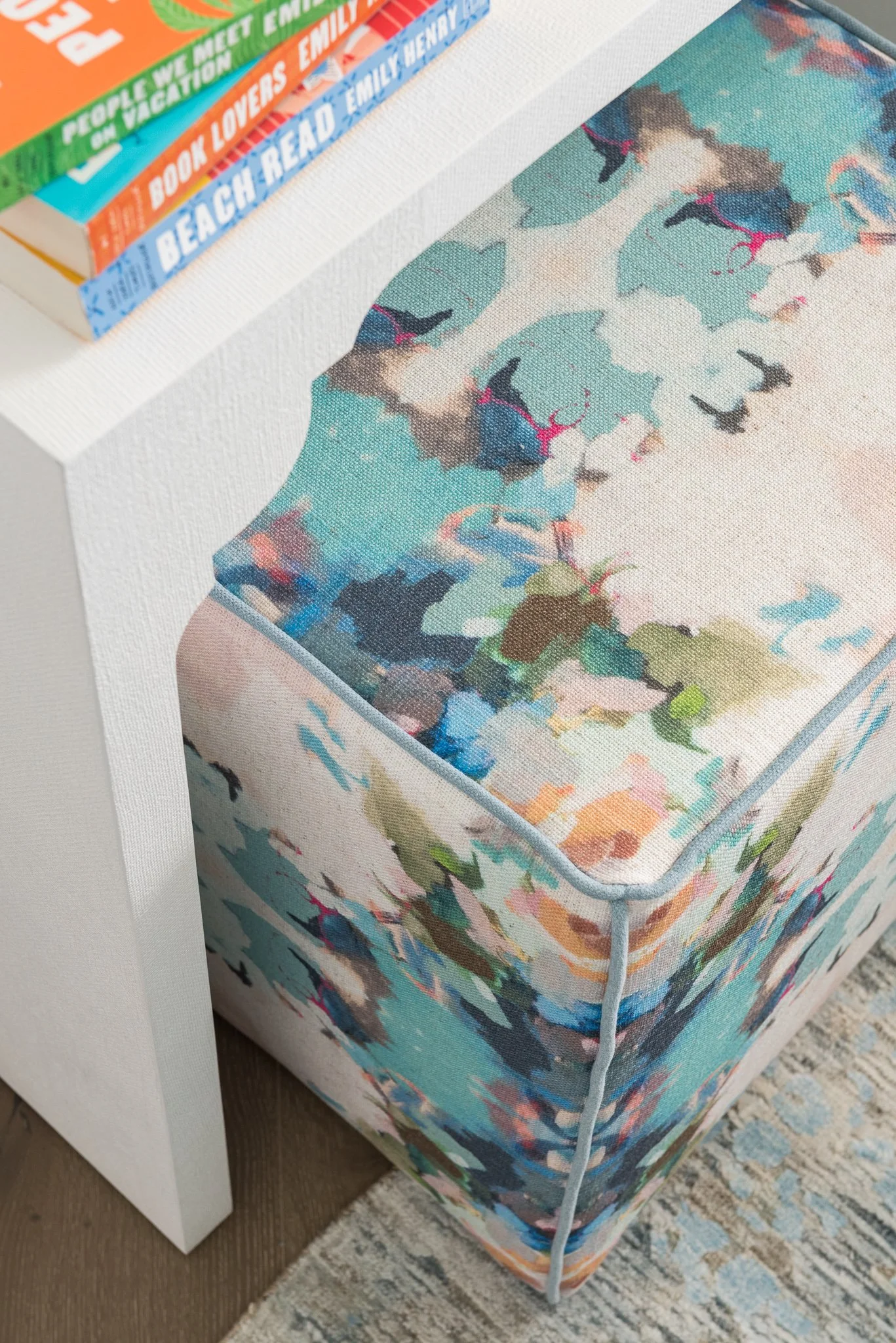 A floral-patterned upholstered ottoman next to a white table with a stack of three colorful books on top.