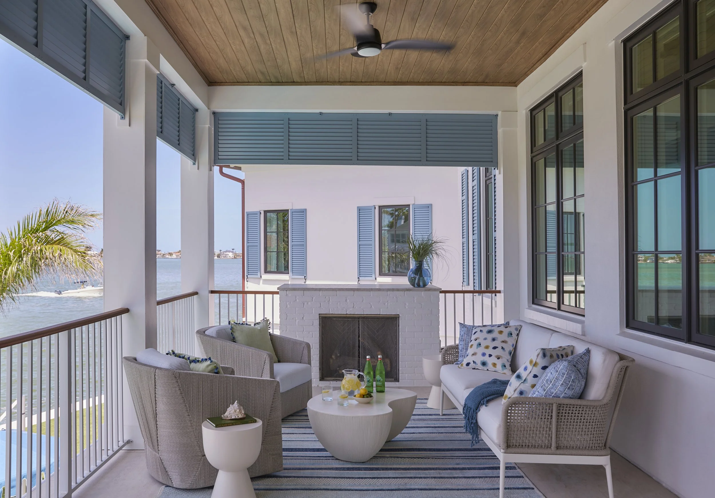 Cozy balcony with white wicker furniture, pillows, a fireplace, and a view of water and palm trees outside.