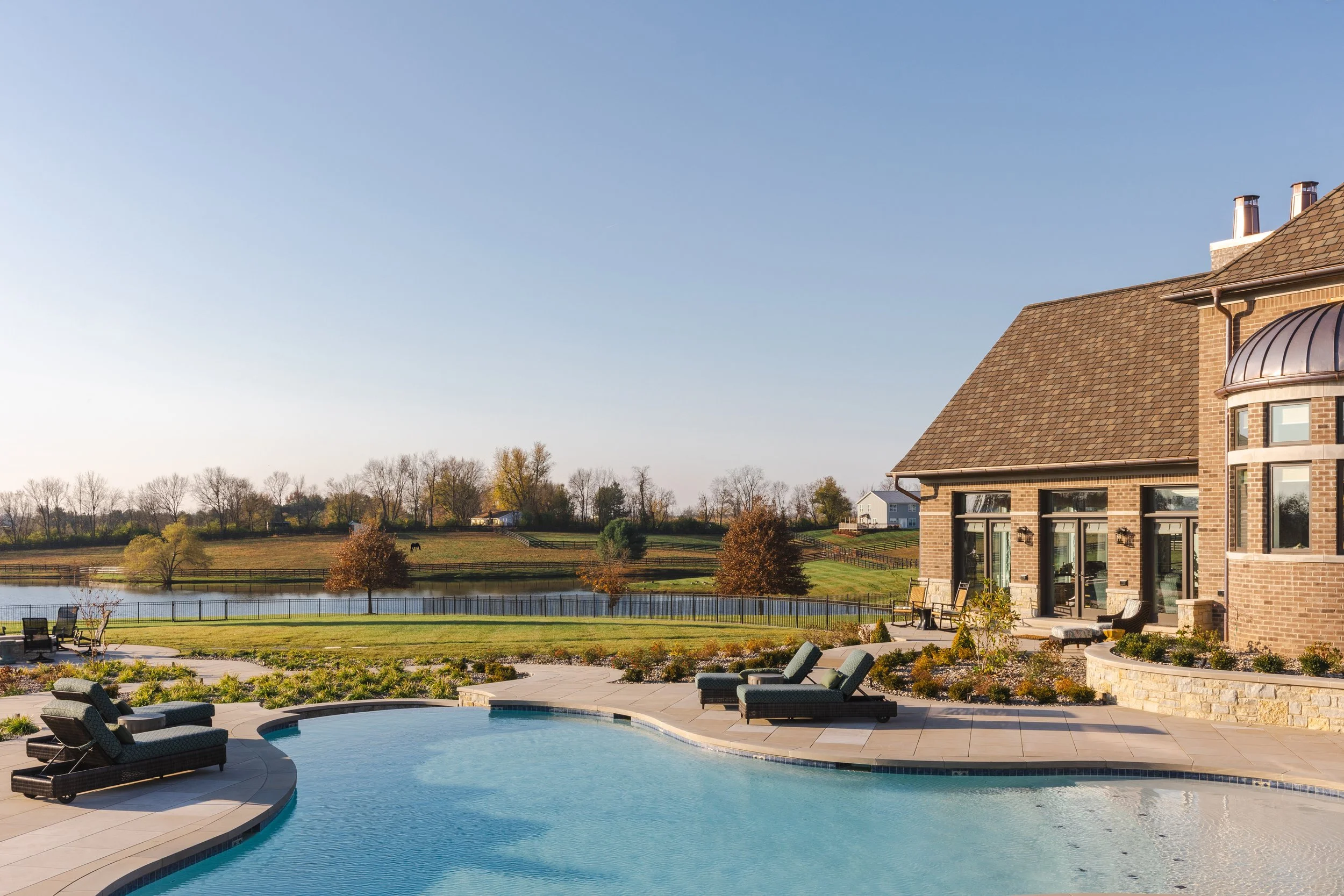 A backyard with a swimming pool, lounge chairs, and a brick house with multiple windows and a curved roof section, overlooking a pond and open fields under a clear blue sky.