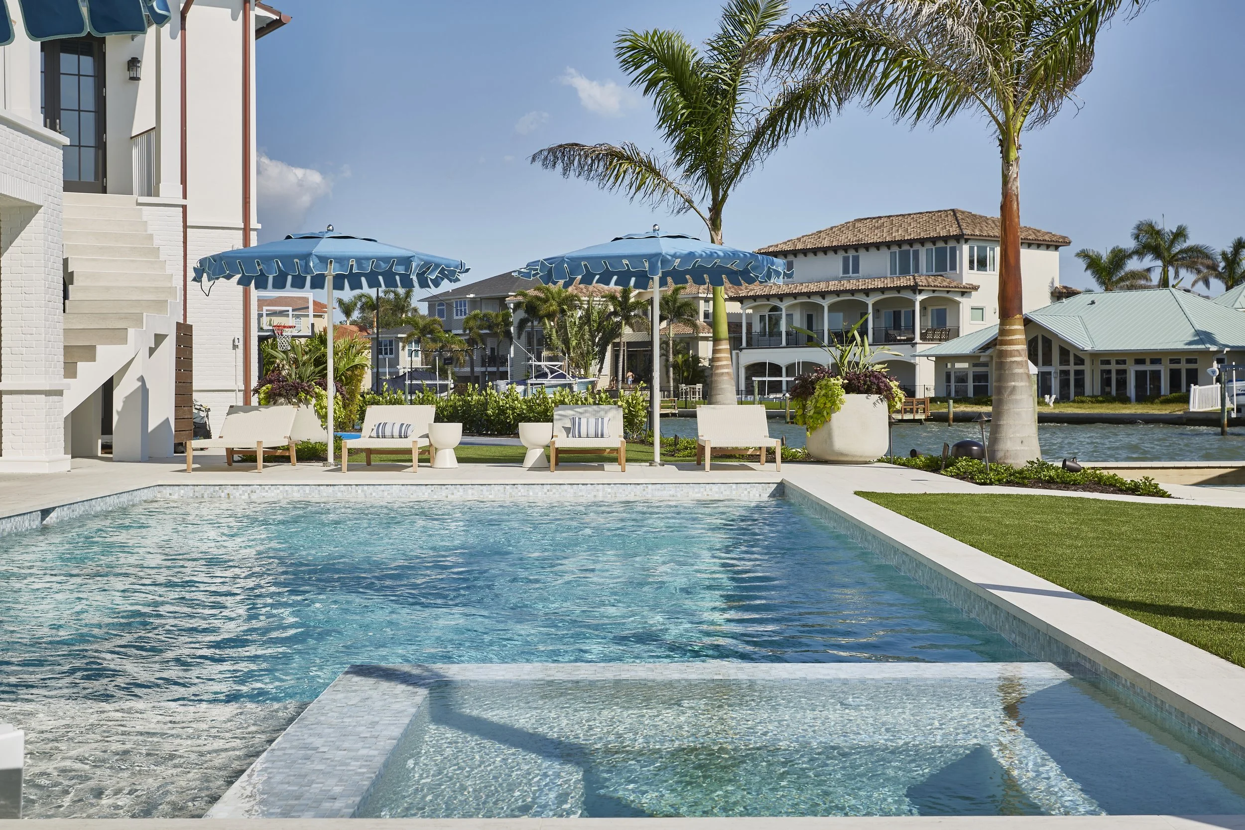 A luxurious outdoor swimming pool area with a hot tub, surrounded by lounge chairs, blue umbrellas, palm trees, and modern houses along a waterway in a sunny and tropical setting.