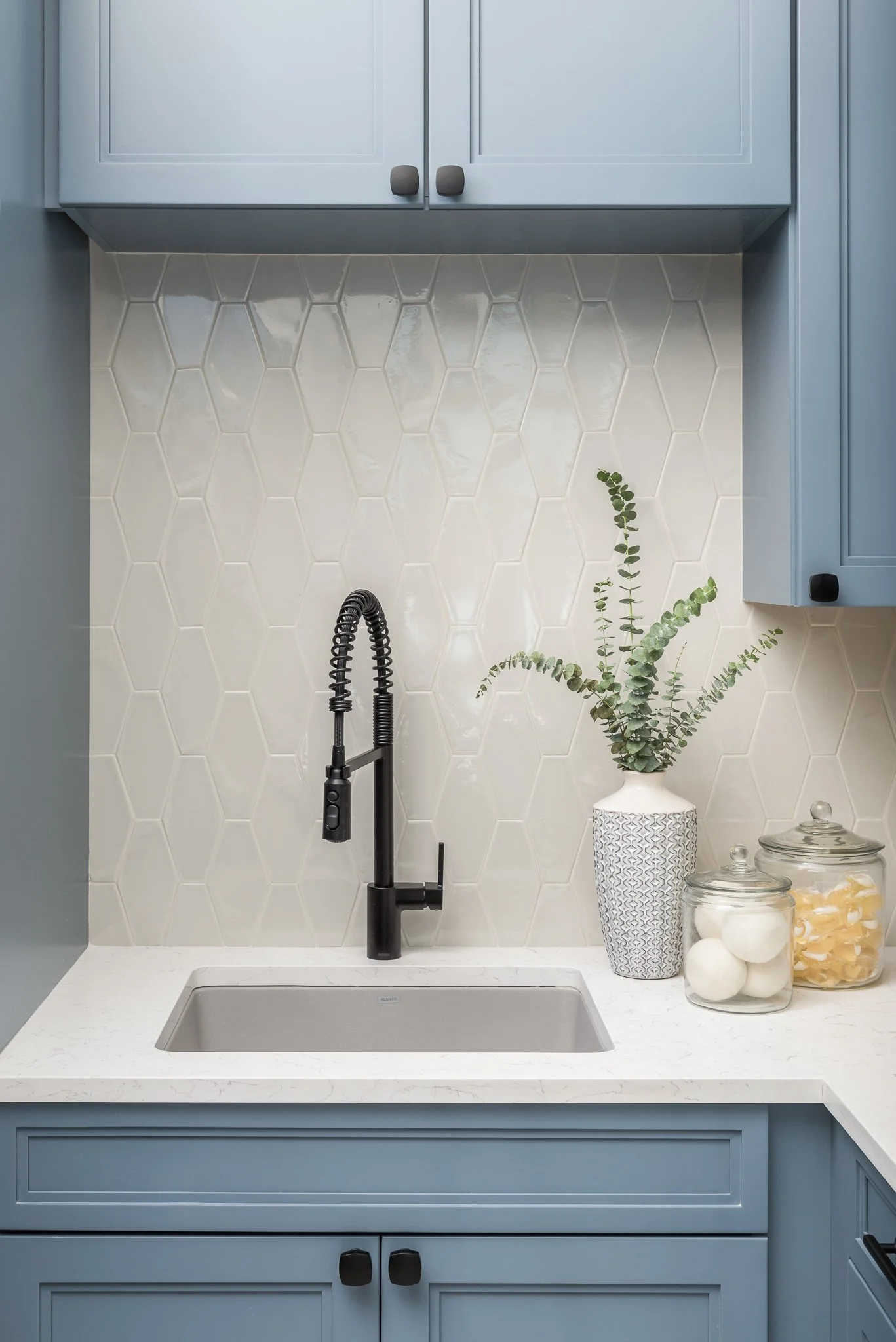 A kitchen sink with a black faucet, white hexagonal tile backsplash, blue cabinets, a white patterned vase with green foliage, and glass jars with decorative items.