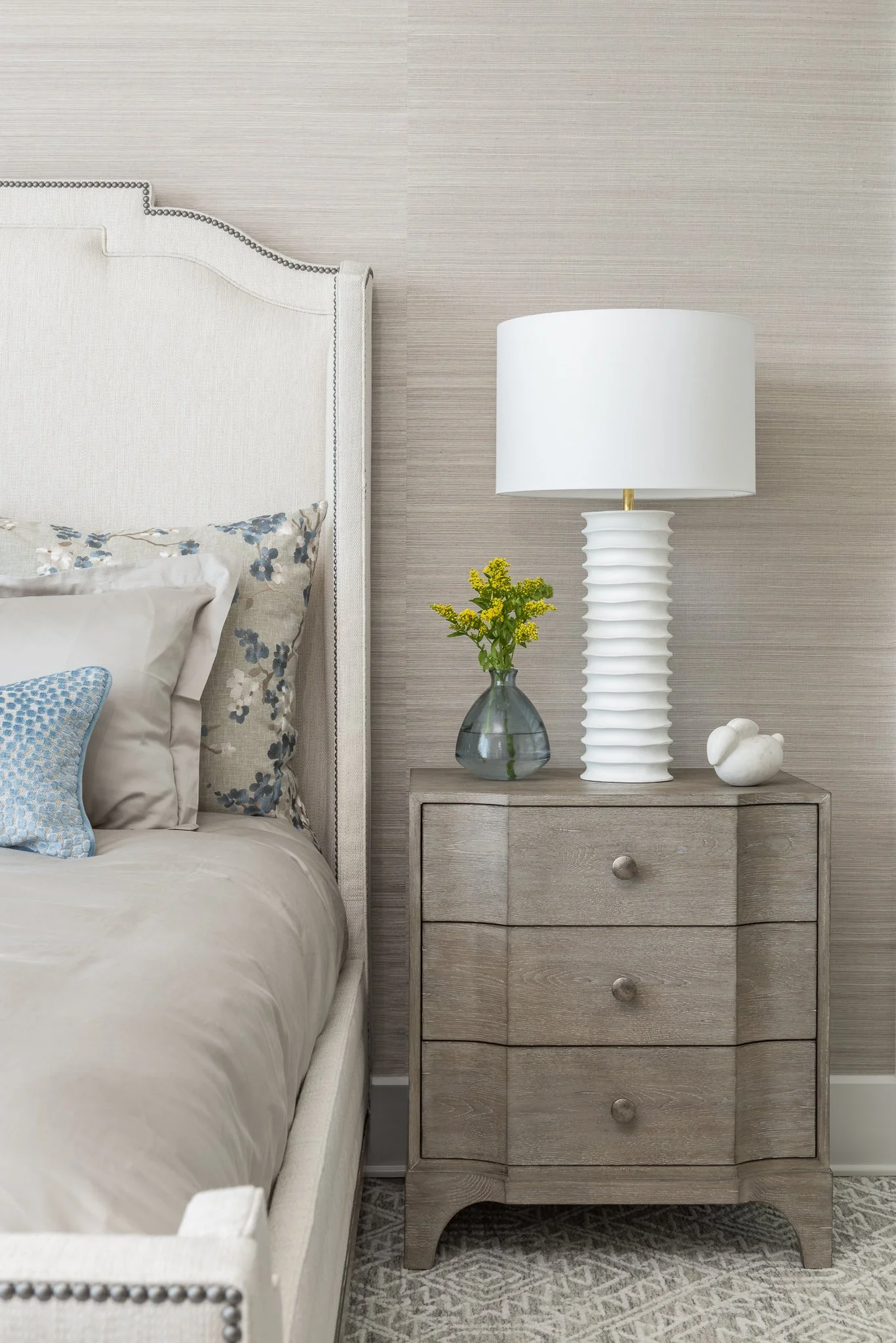 A cozy bedroom corner featuring a white upholstered headboard, a beige bedding set with pillows, a light wood nightstand with three drawers, a white textured table lamp, a small glass vase with yellow flowers, and a small white ceramic sculpture. The