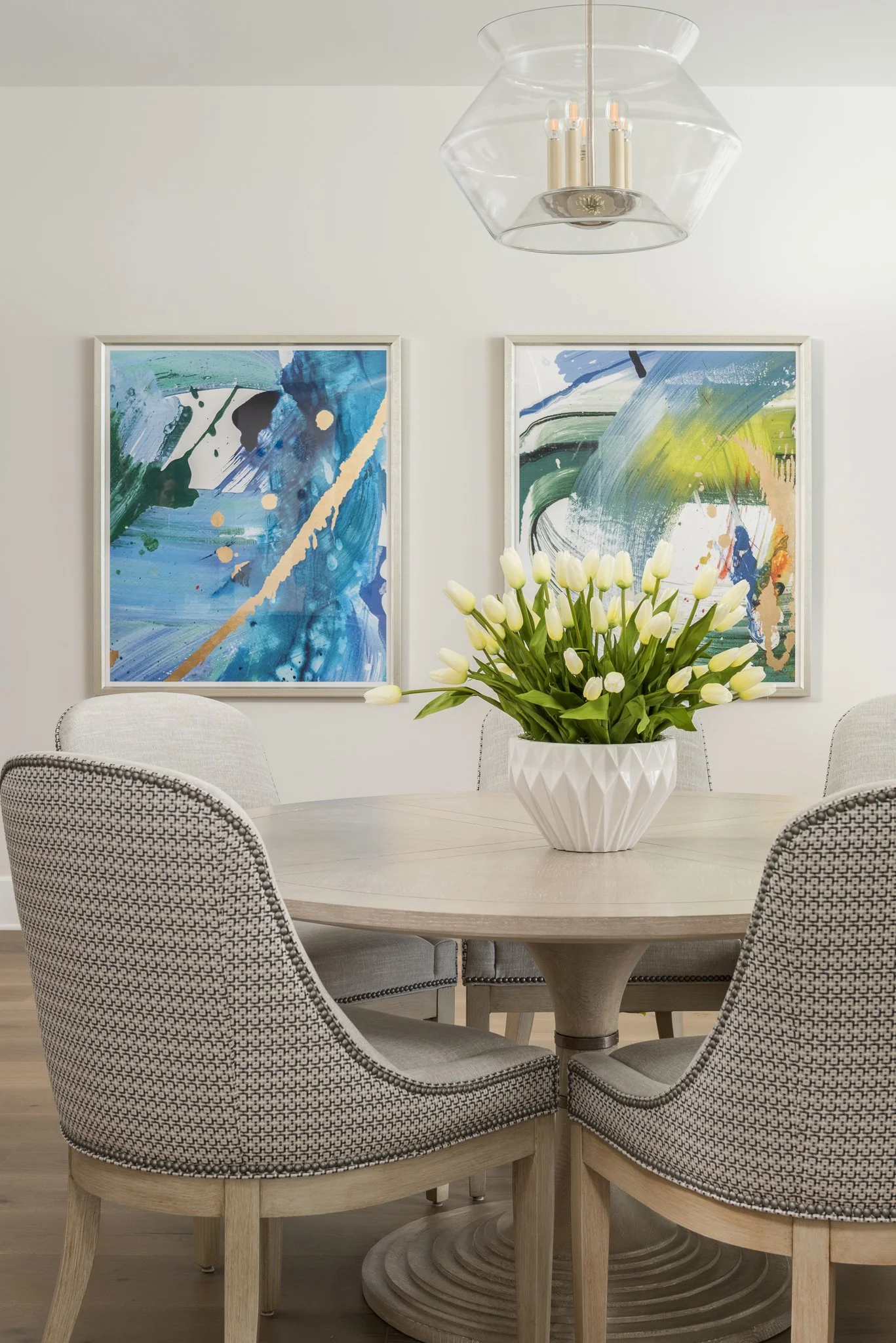 A round wooden dining table with a white geometric-patterned vase of white tulips in the center. Four upholstered dining chairs surround the table. On the wall behind, there are two abstract paintings in white frames, and a modern chandelier hangs ab