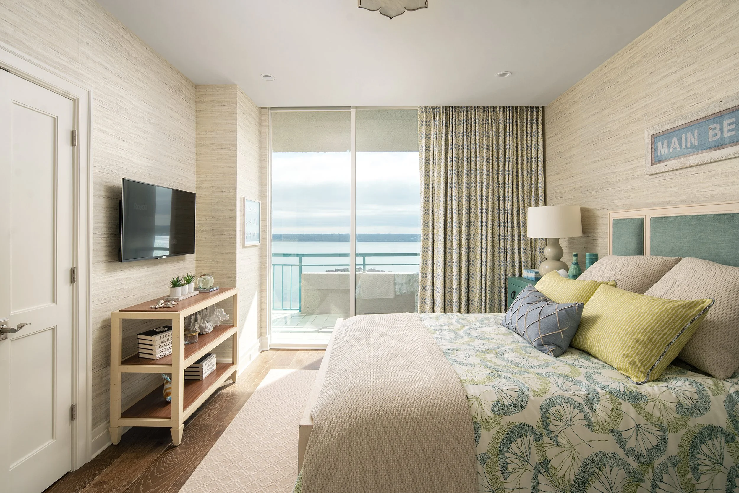 A bedroom with a sliding glass door leading to a balcony overlooking a body of water. The bed has a floral patterned bedspread with green and blue pillows. There is a nightstand with a lamp and decorative items. A TV is mounted on the wall opposite t
