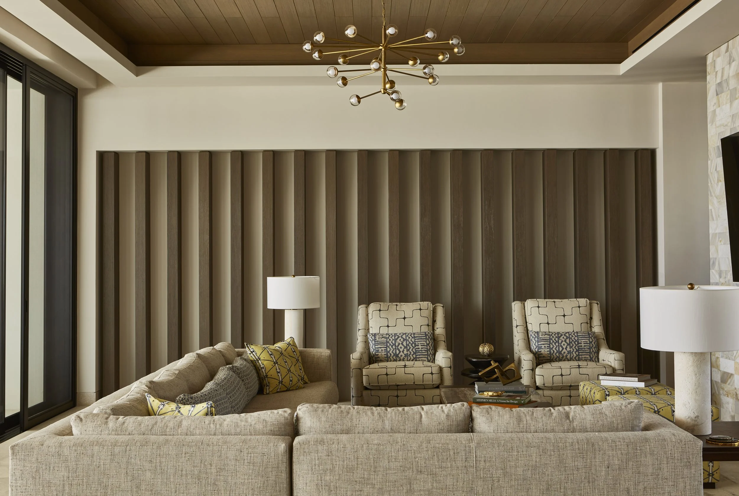 Modern living room with beige sofa, two matching armchairs, side tables with books and decorative objects, lamps, wood-paneled wall, and a metal chandelier ceiling light.