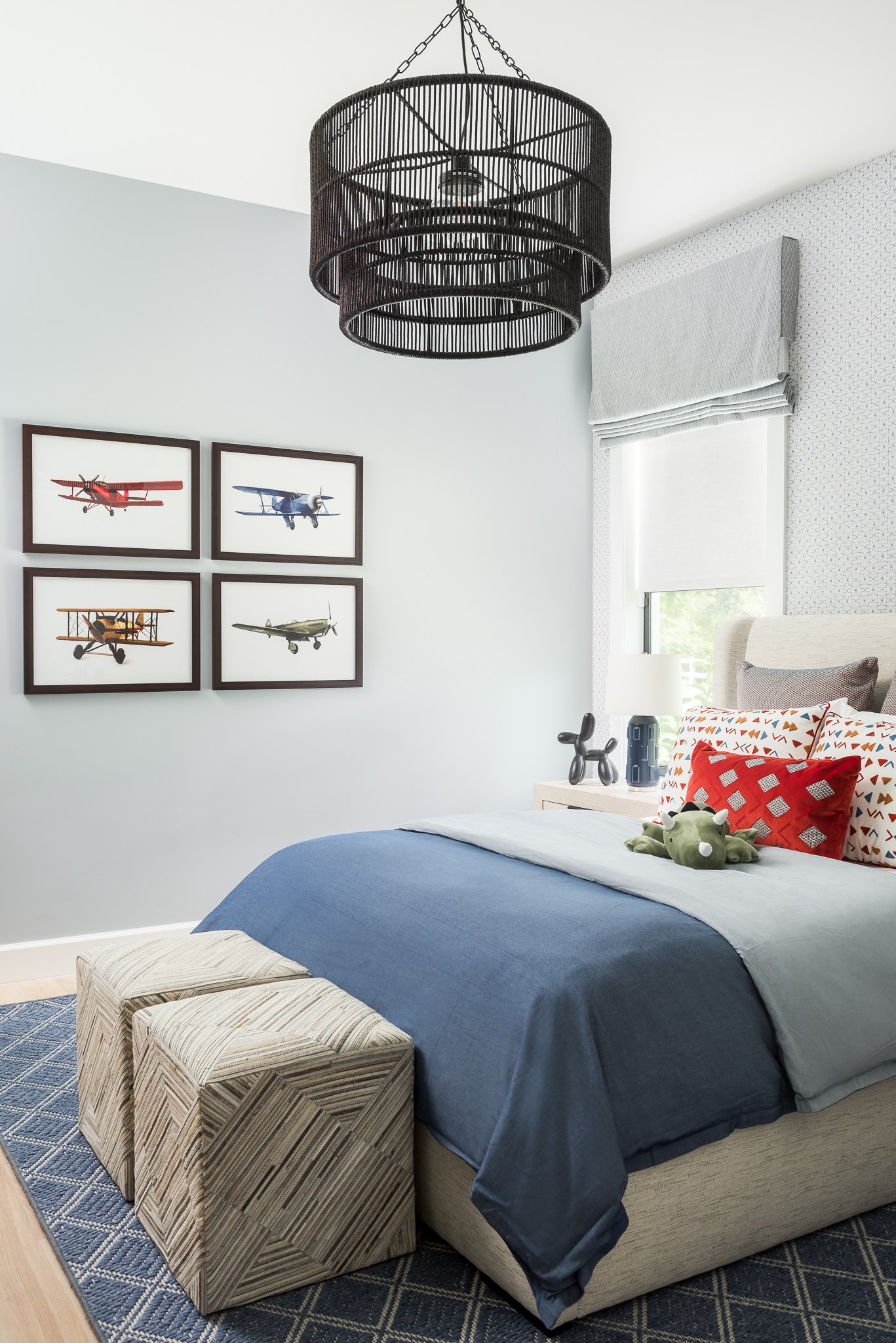 A bedroom with a bed covered in blue and gray bedding, decorated with pillows and a stuffed toy, with framed airplane pictures on the wall and a black chandelier hanging from the ceiling.