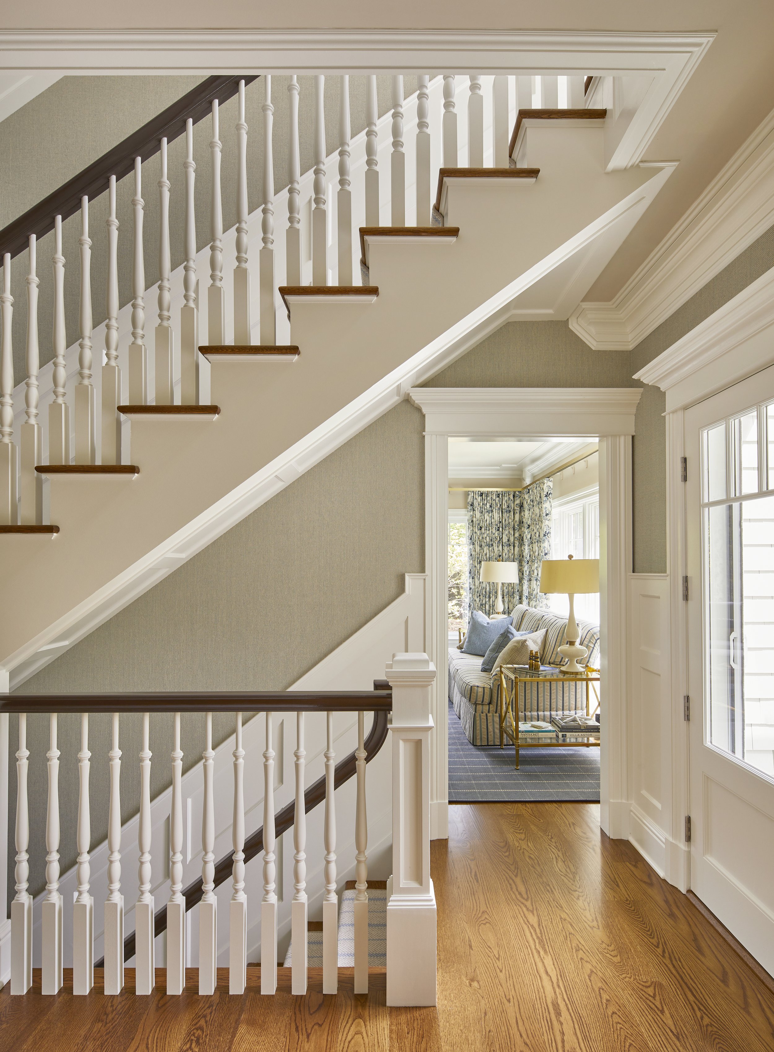 Interior view of a house with a staircase featuring white spindles and brown handrail, leading to upper floor, with a cozy living room visible through an open doorway, decorated with a sofa, cushions, lamps, and floral curtains.