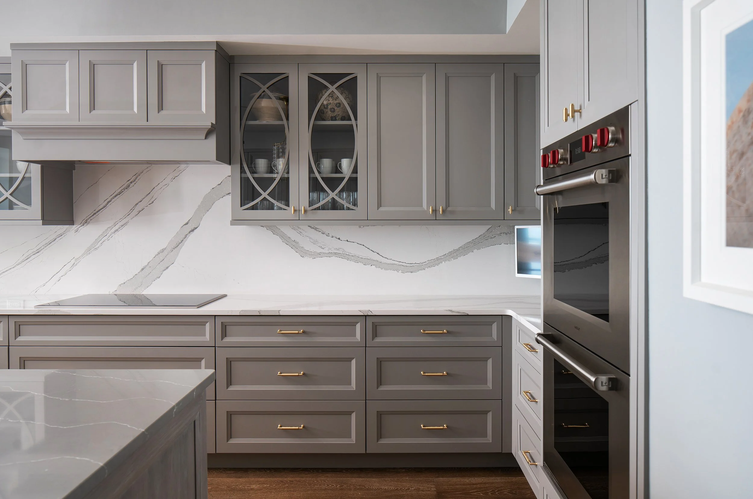 Modern kitchen with grey cabinets, gold handles, marble countertop, and a built-in double oven.