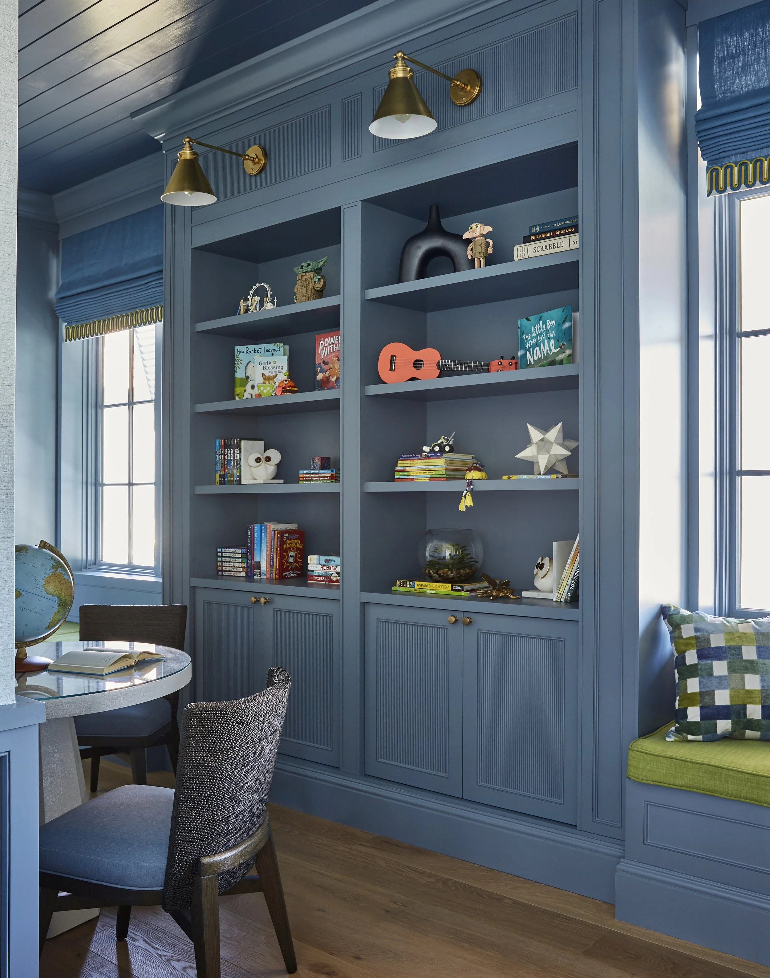 A blue built-in bookshelf with decorative and children's items, a window with blue window valances, and part of a round table with chairs in a room with hardwood floors.
