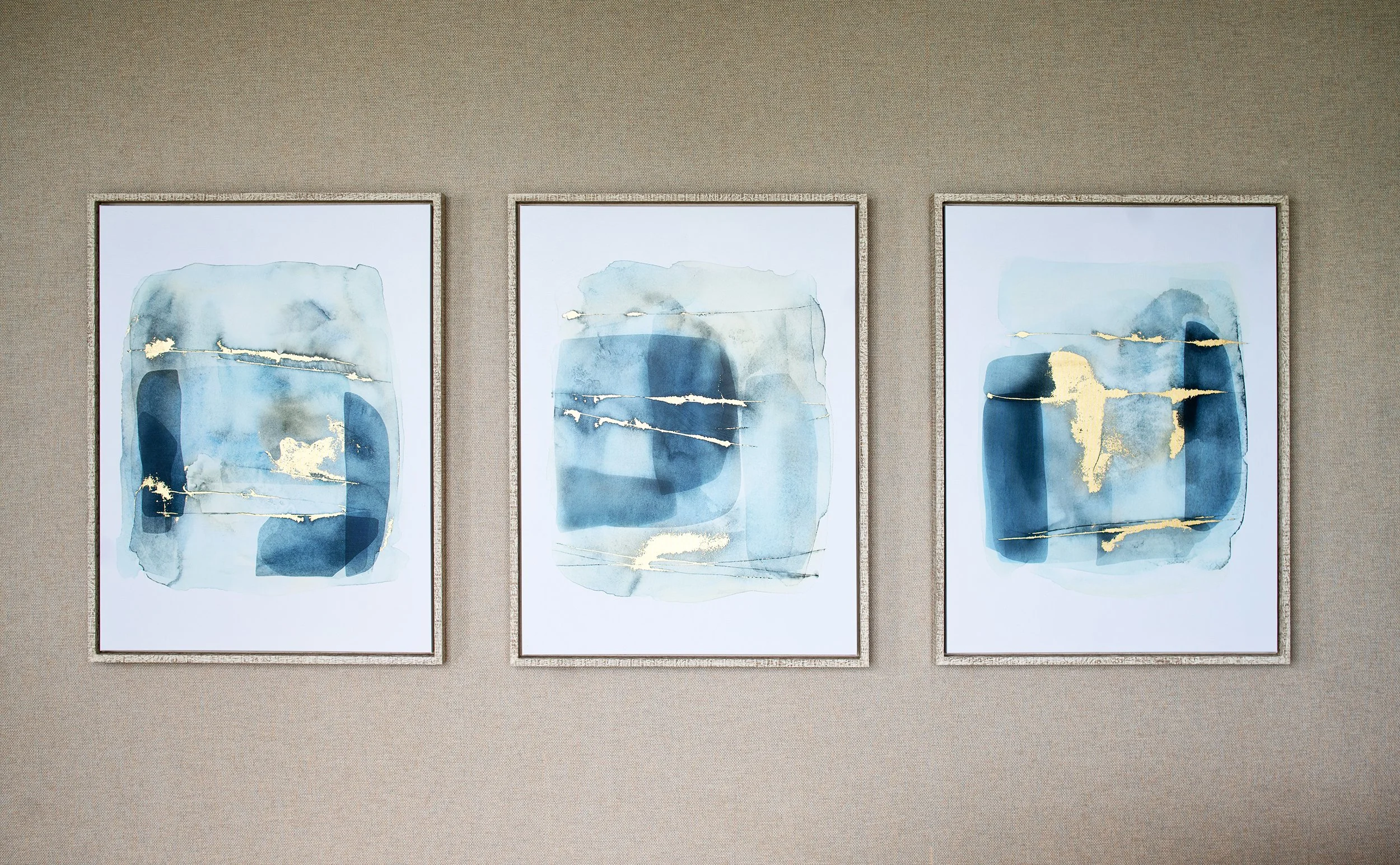 Three abstract watercolor paintings in cool blue tones with gold accents, framed and displayed on a beige wall.