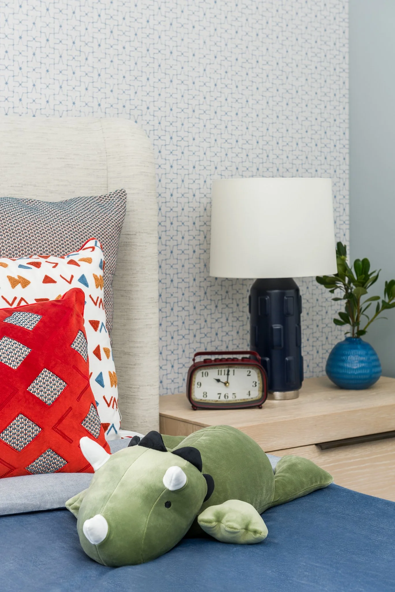 Close-up of a bedroom nightstand with a vintage alarm clock, a modern table lamp, and a blue vase with a green plant. Part of a bed with decorative pillows and a plush dinosaur toy are visible in the foreground, with patterned wallpaper in the backgr