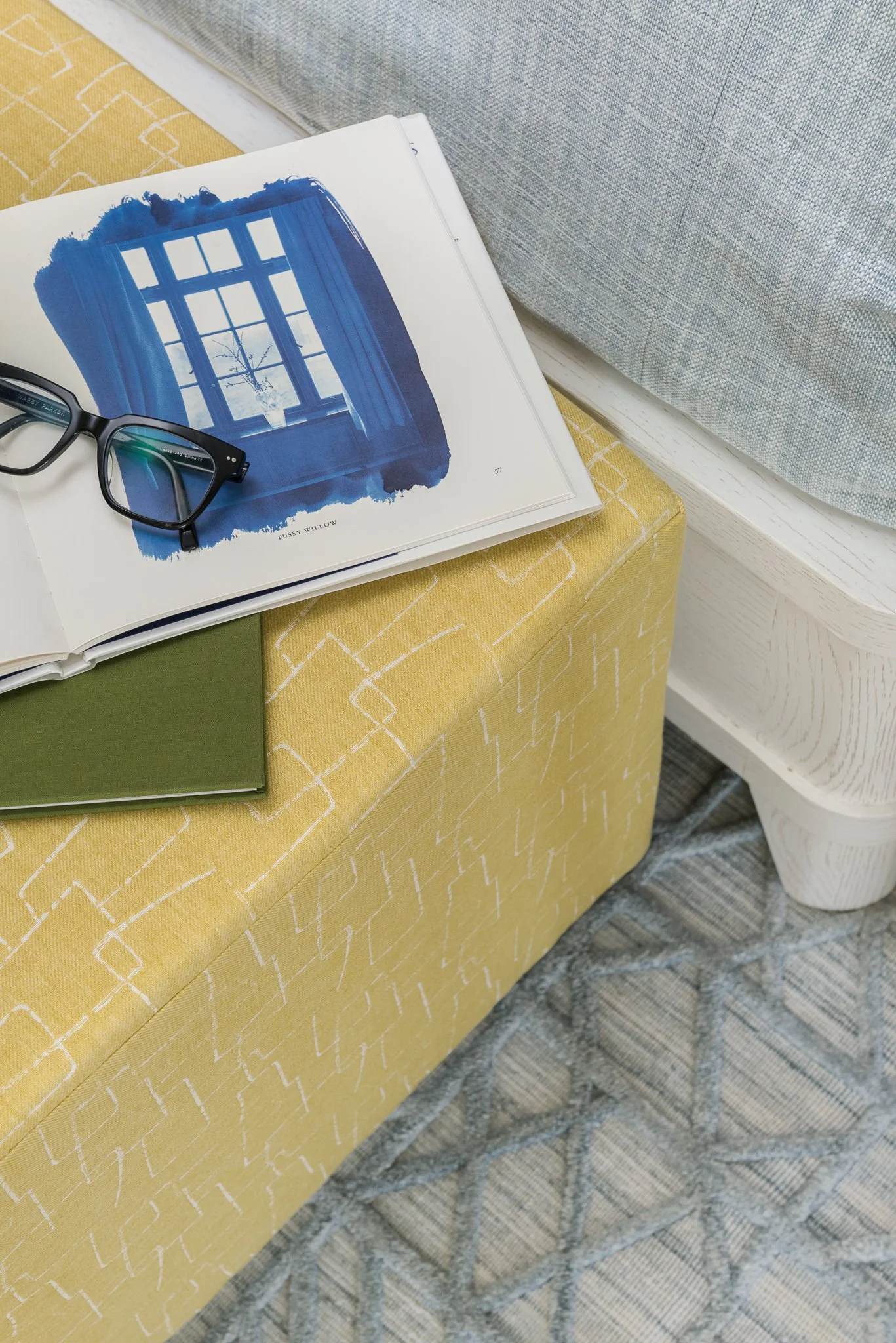 An open book with a blue watercolor painting of a window and curtains, a pair of black eyeglasses resting on the page, a closed green book next to it, a yellow patterned upholstered bench, a white wooden side table, a gray and white patterned rug on 