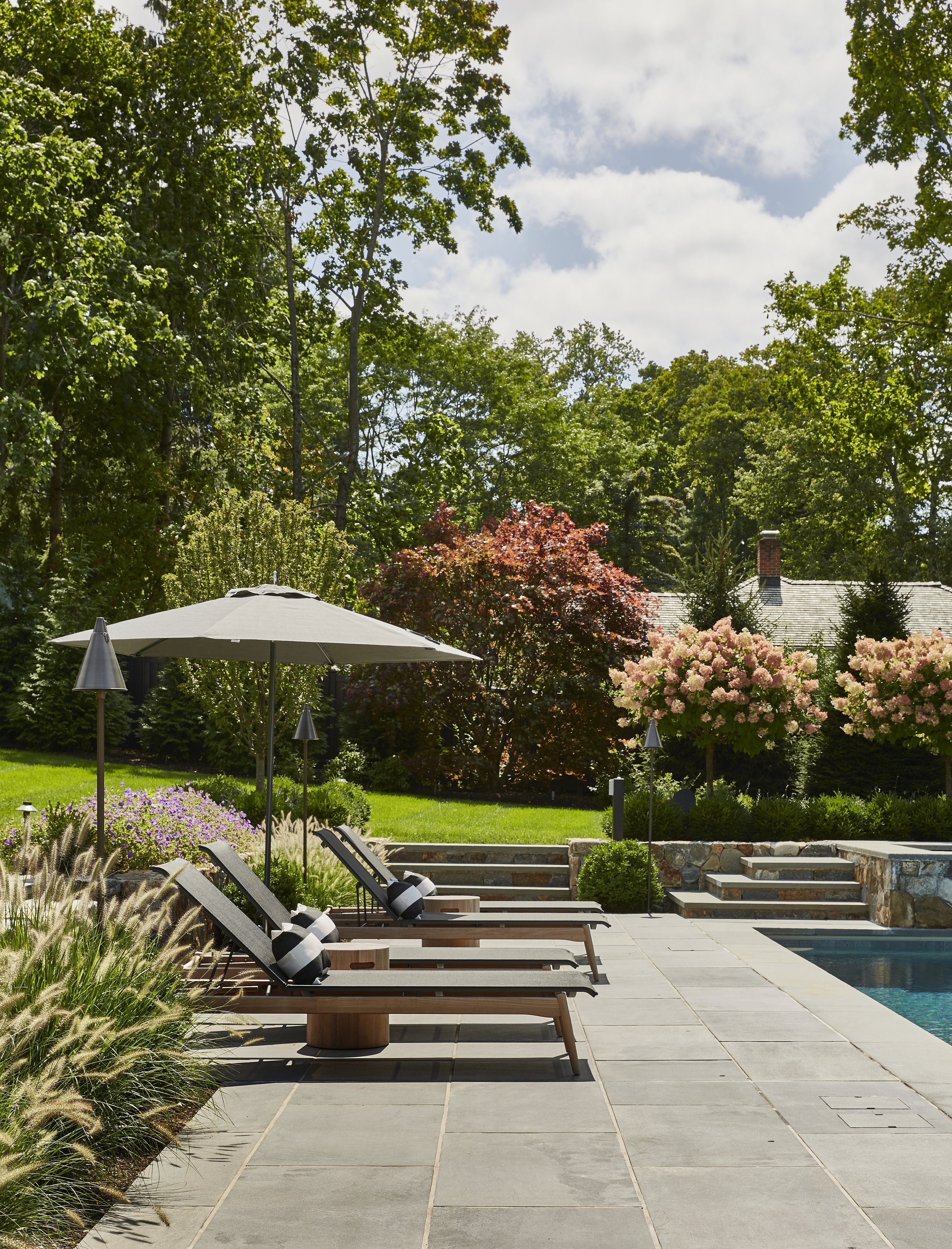 A backyard with poolside lounge chairs under an umbrella, surrounded by trees, flowering bushes, and a house in the background on a sunny day.