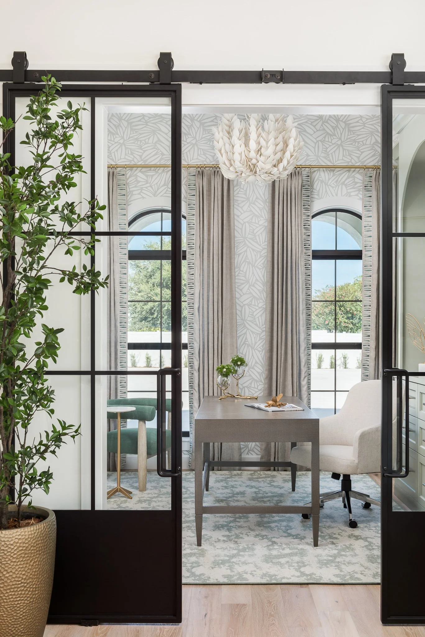 View of a modern office with sliding glass doors, large windows, beige curtains, a chandelier, a desk with decorative items, a white upholstered chair, and a branching plant in a textured planter.