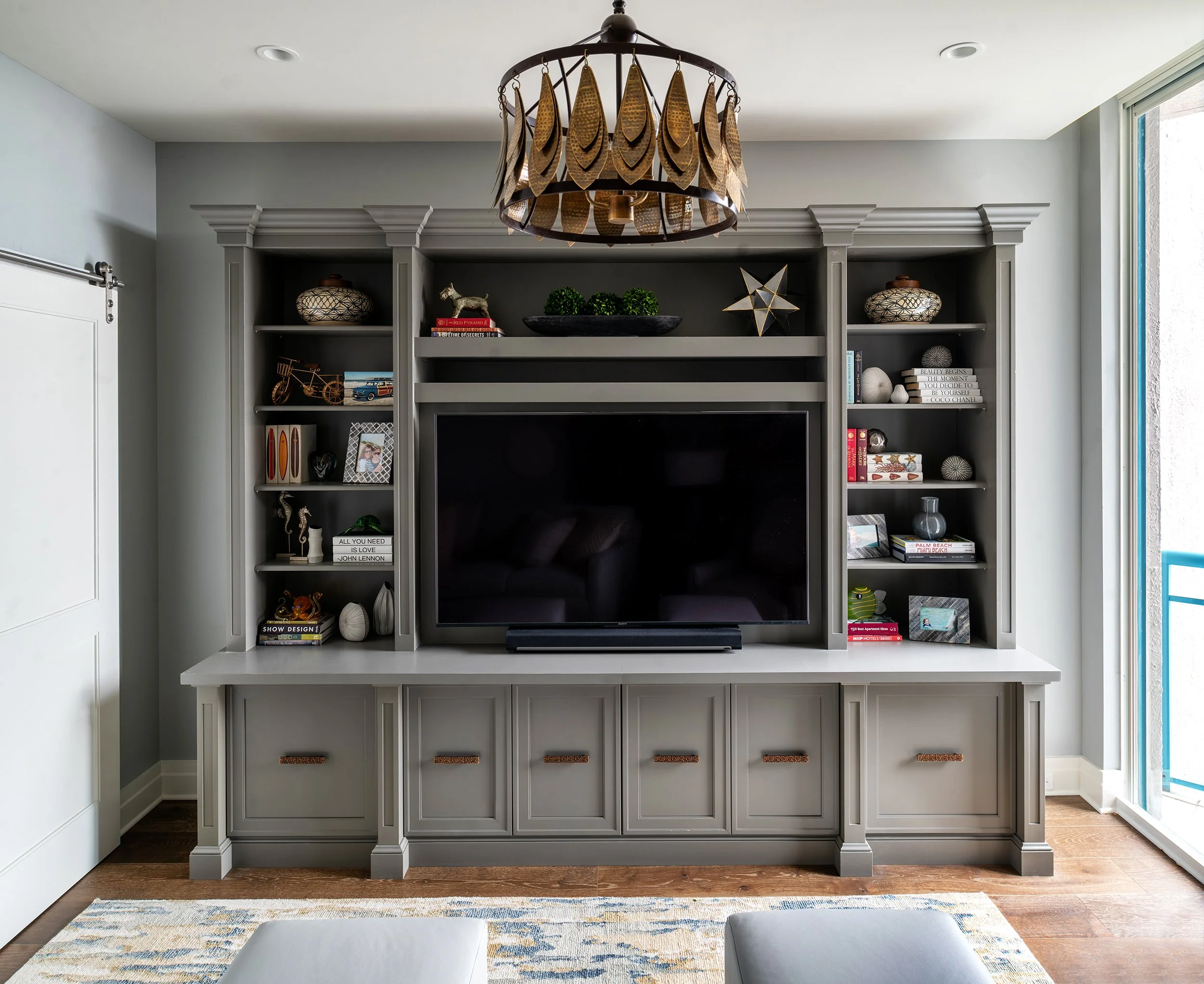 Living room TV entertainment center with grey built-in shelves and cabinets, decorated with books, vases, picture frames, and decorative objects, with a large flat-screen TV in the center, a pendant light hanging from the ceiling, hardwood floors, an