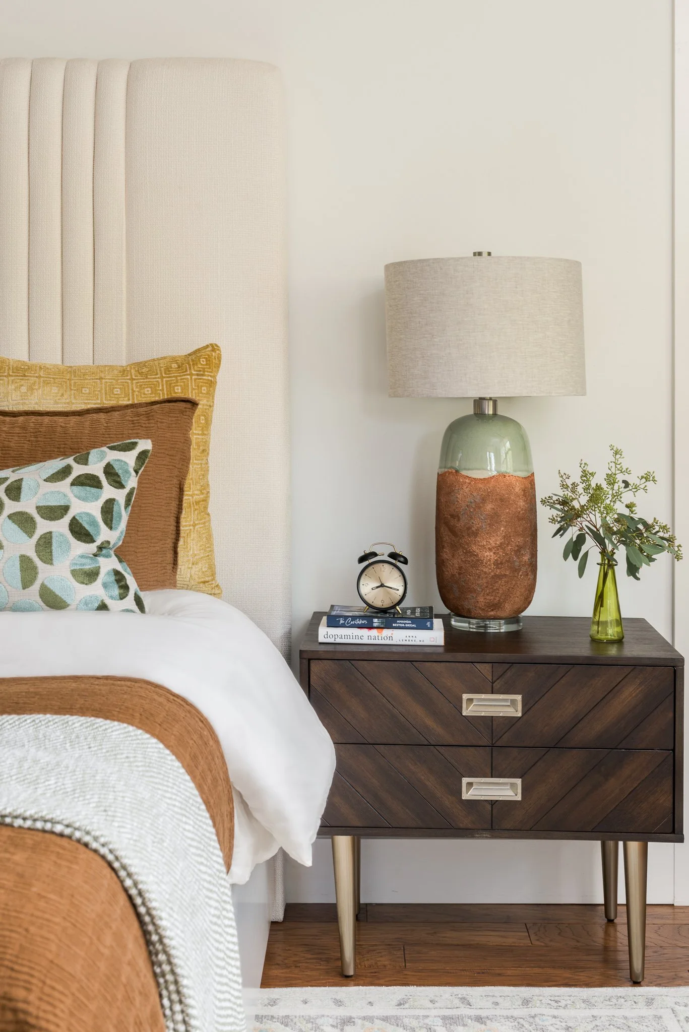 A bedroom with a nightstand holding a ceramic lamp, an alarm clock, and a small green vase with greenery, next to a bed with colorful pillows.