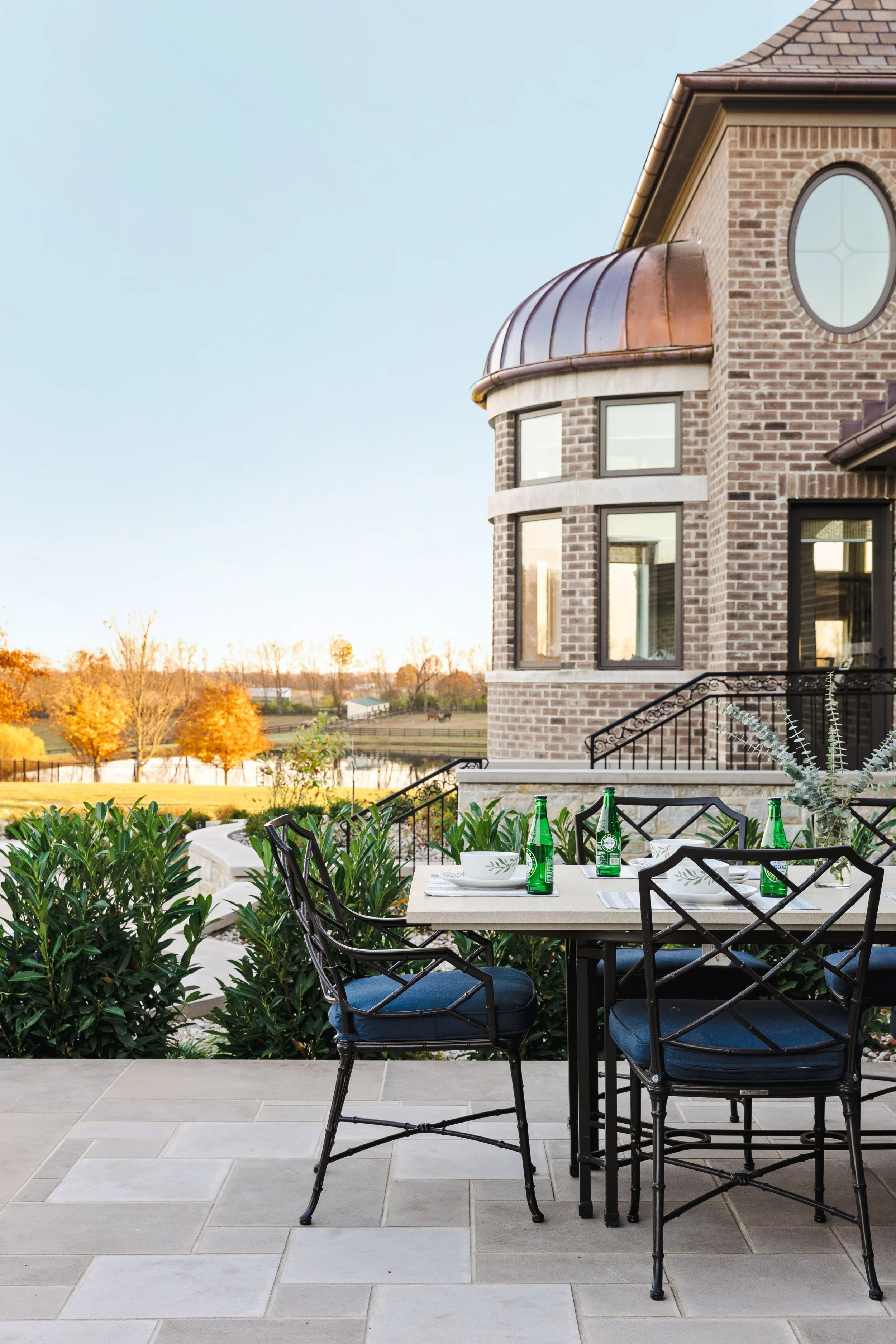 An outdoor dining area on a patio with a table set with plates and bottles, surrounded by greenery, in front of a brick house with large windows and a copper roof, overlooking a field with trees and a pond in the background during sunset.