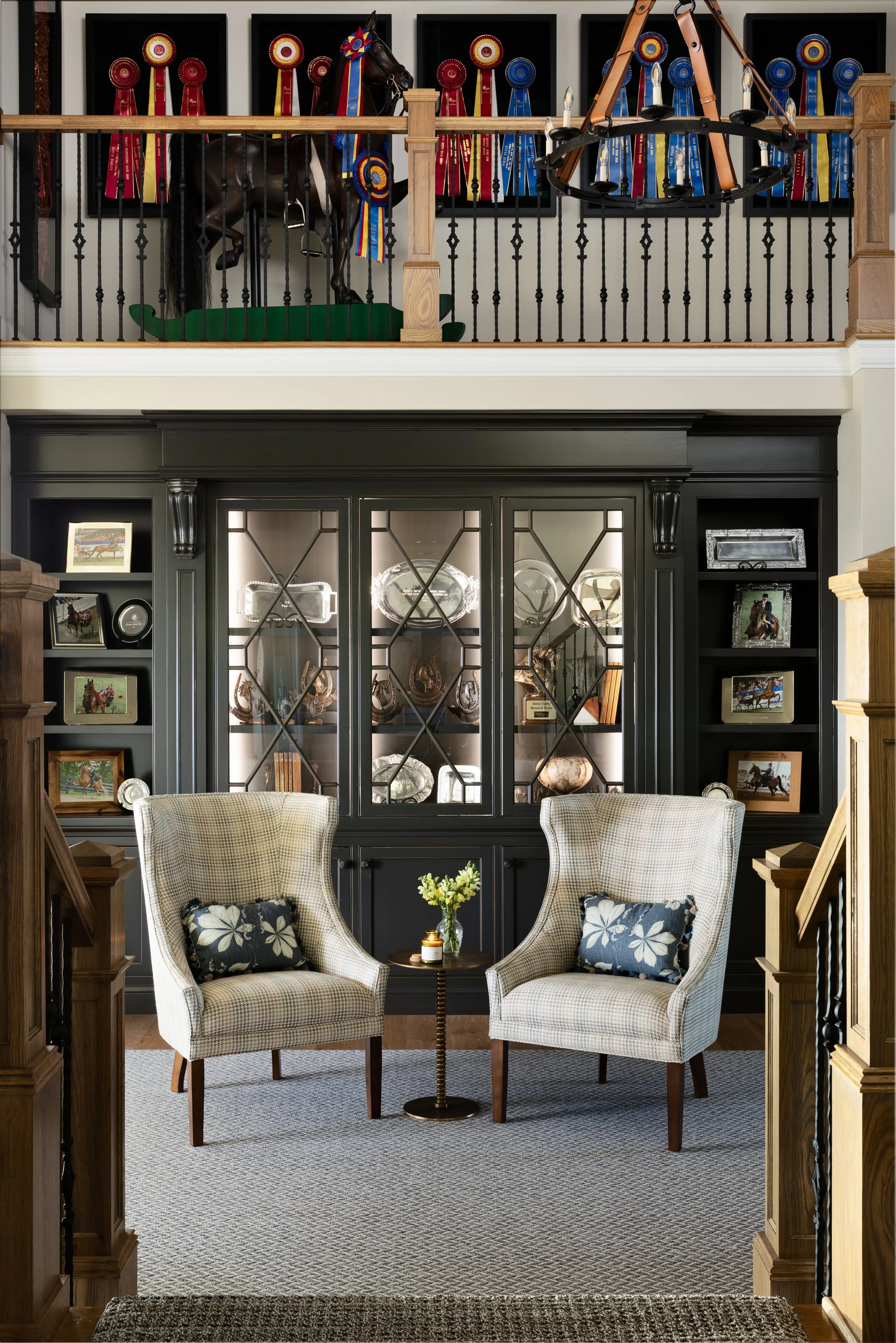A cozy living room with two armchairs, a small table with a flower vase, and a black display cabinet with glass doors, flanked by shelves showing photos and collectibles. The upper level features a horse figurine with multiple ribbons and rosettes in