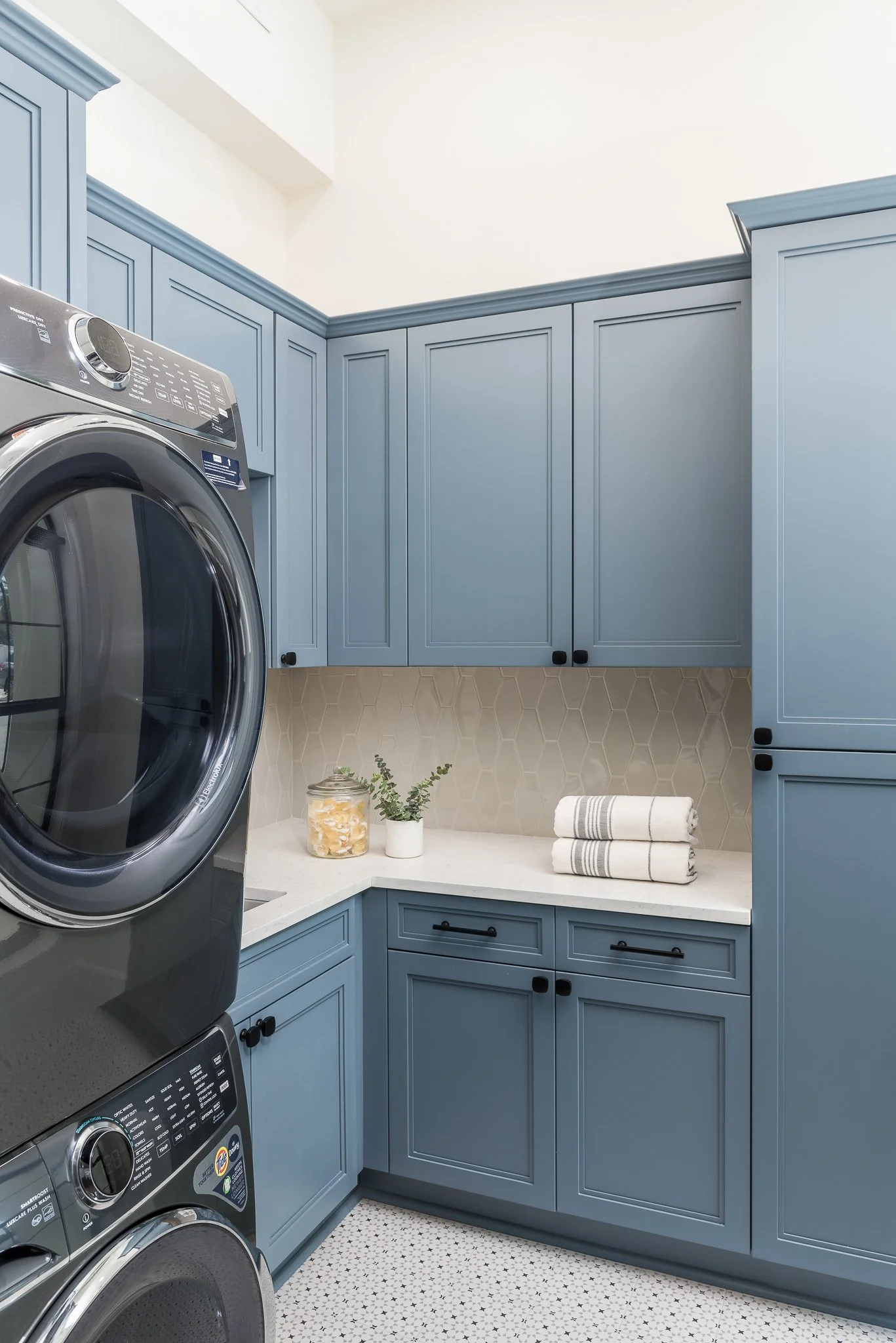 Laundry room with blue cabinets, stacked washer and dryer, beige hexagonal backsplash, white countertop, decorative jars, and folded towels.