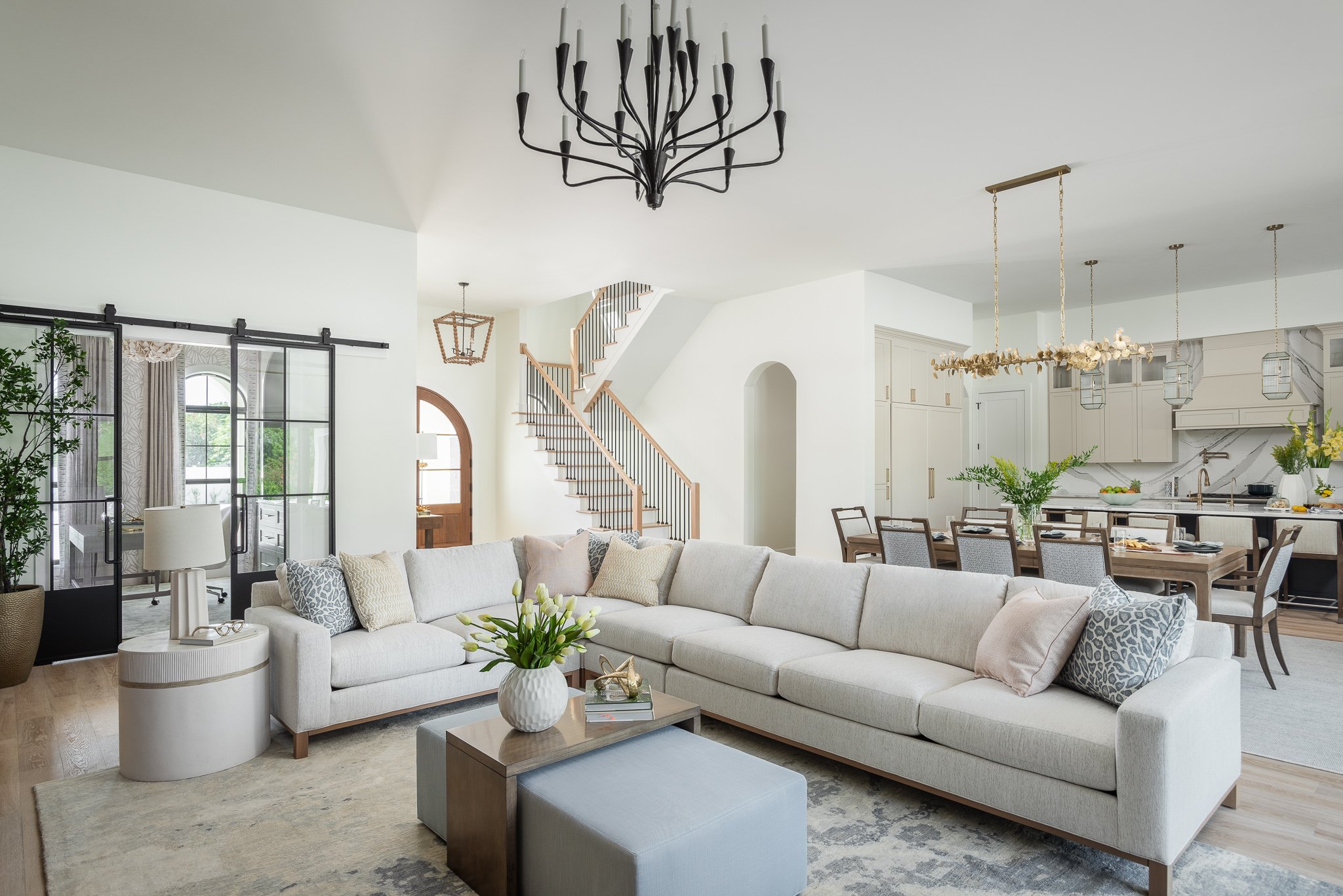 Spacious and modern open-concept living room with a large white sectional sofa, coffee table with a vase of white tulips, and a neutral-toned area rug. Behind the sofa, there's a dining area with a wooden table and gray chairs, and a bright kitchen w