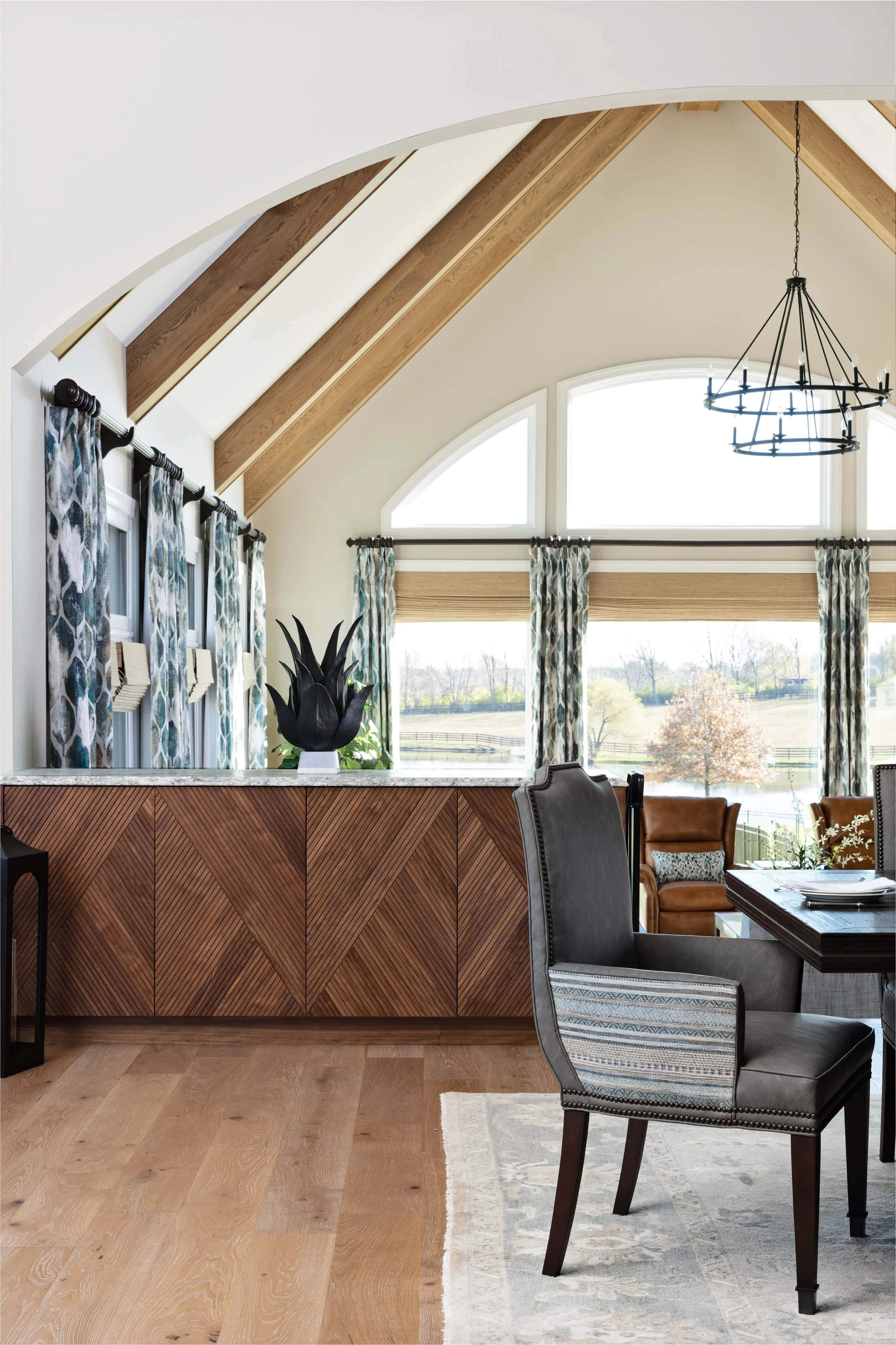 A spacious dining room with high vaulted ceiling and exposed wooden beams, large windows with patterned curtains, a chandelier, and modern furniture including a black padded chair and a wooden table.